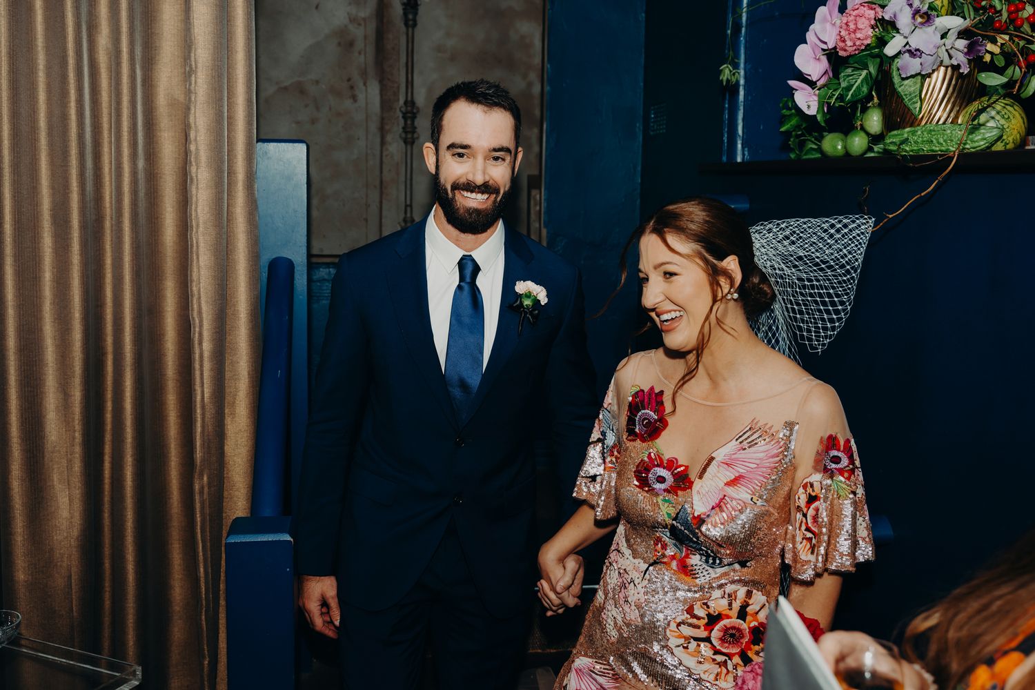 A couple in formal attire poses elegantly against a backdrop with floral arrangements.
