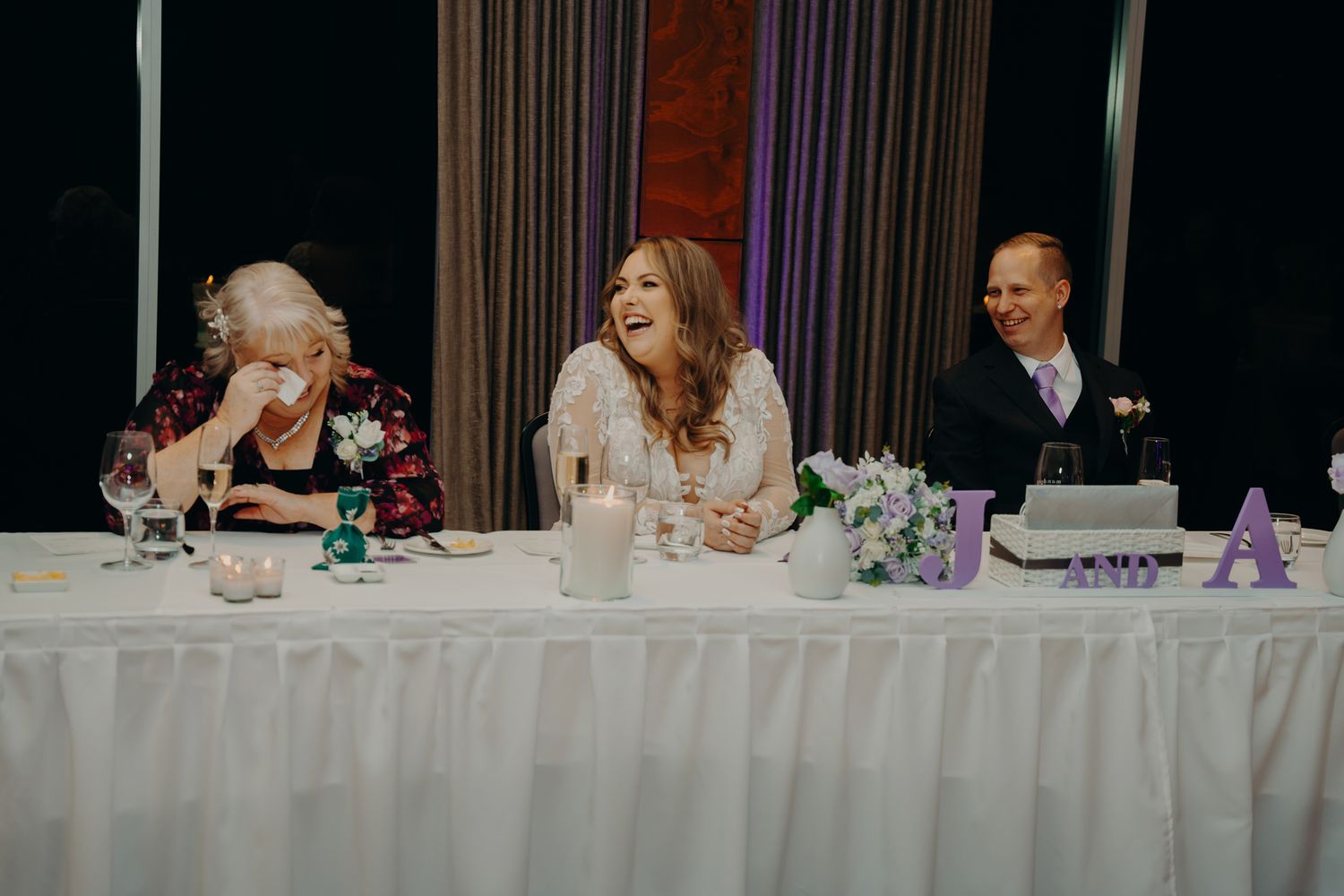Wedding party seated at an elegant head table with purple floral decorations and white linens.