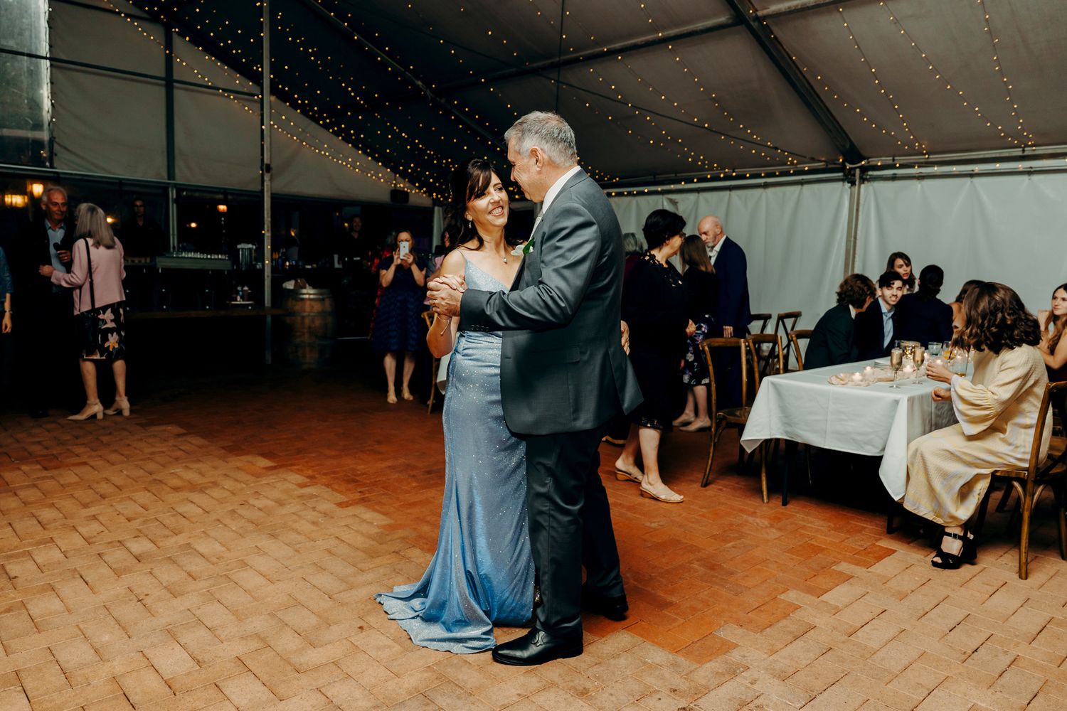 A couple in formal attire share an intimate dance on a wooden floor under a tent at an evening celebration.