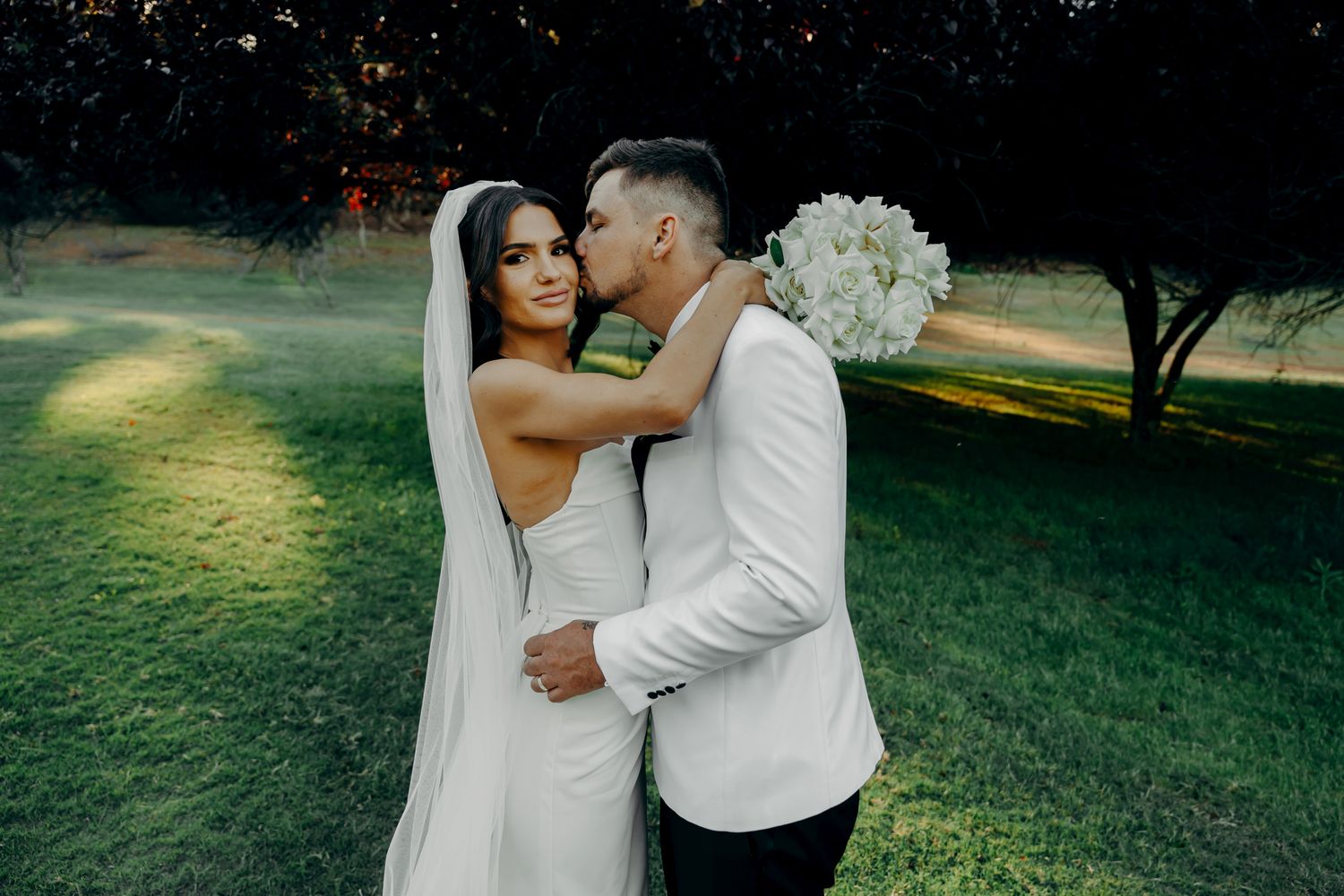 A romantic couple in white wedding attire embraces in a sunlit garden setting during their special day.