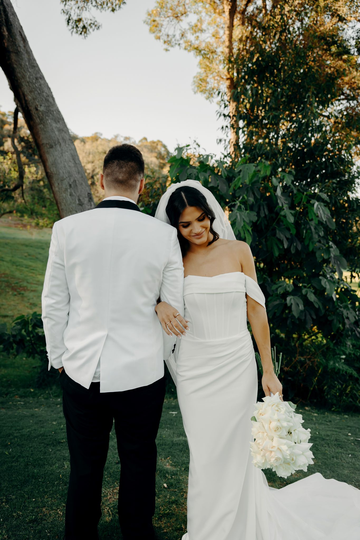 A romantic wedding couple embraces outdoors with the bride holding a white bouquet against an autumn landscape.