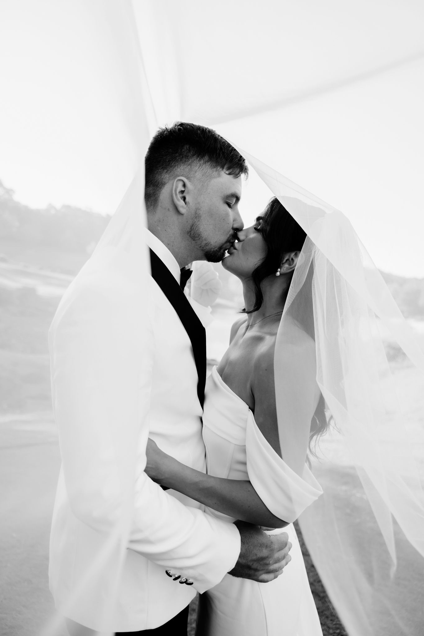 A romantic black and white wedding photo showing a tender moment between newlyweds under a flowing veil.