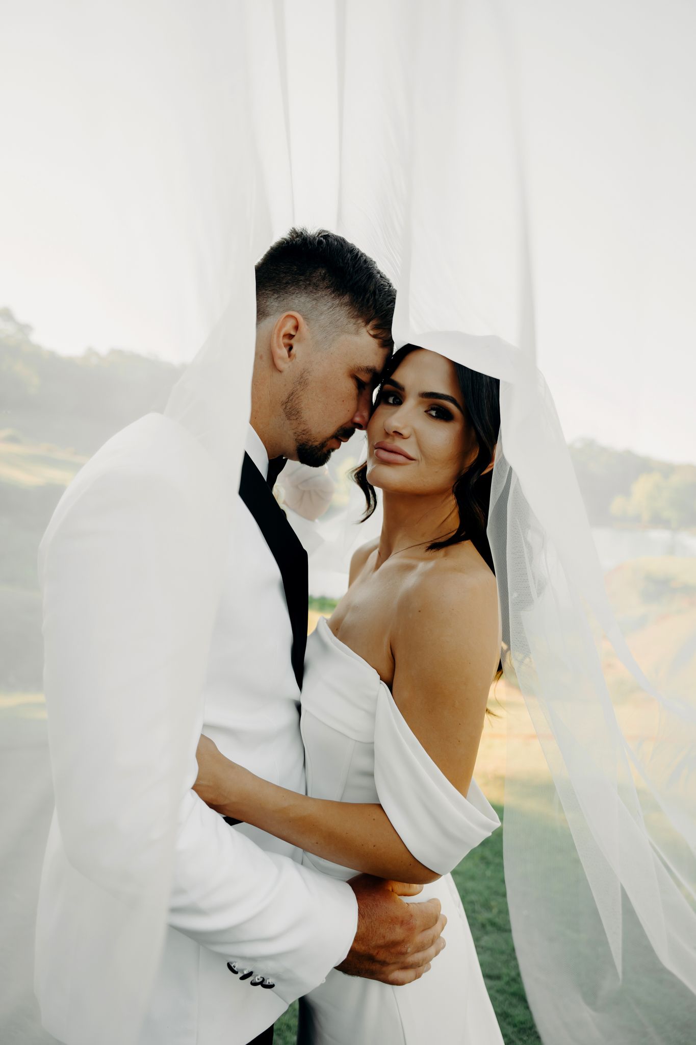 Intimate wedding portrait of a couple in white attire sharing a tender moment against a bright background.