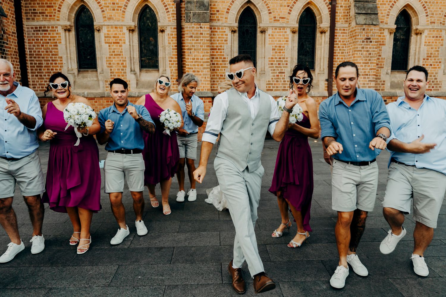 Wedding party celebrates with drinks outside gothic stone church with arched windows during summer celebration.