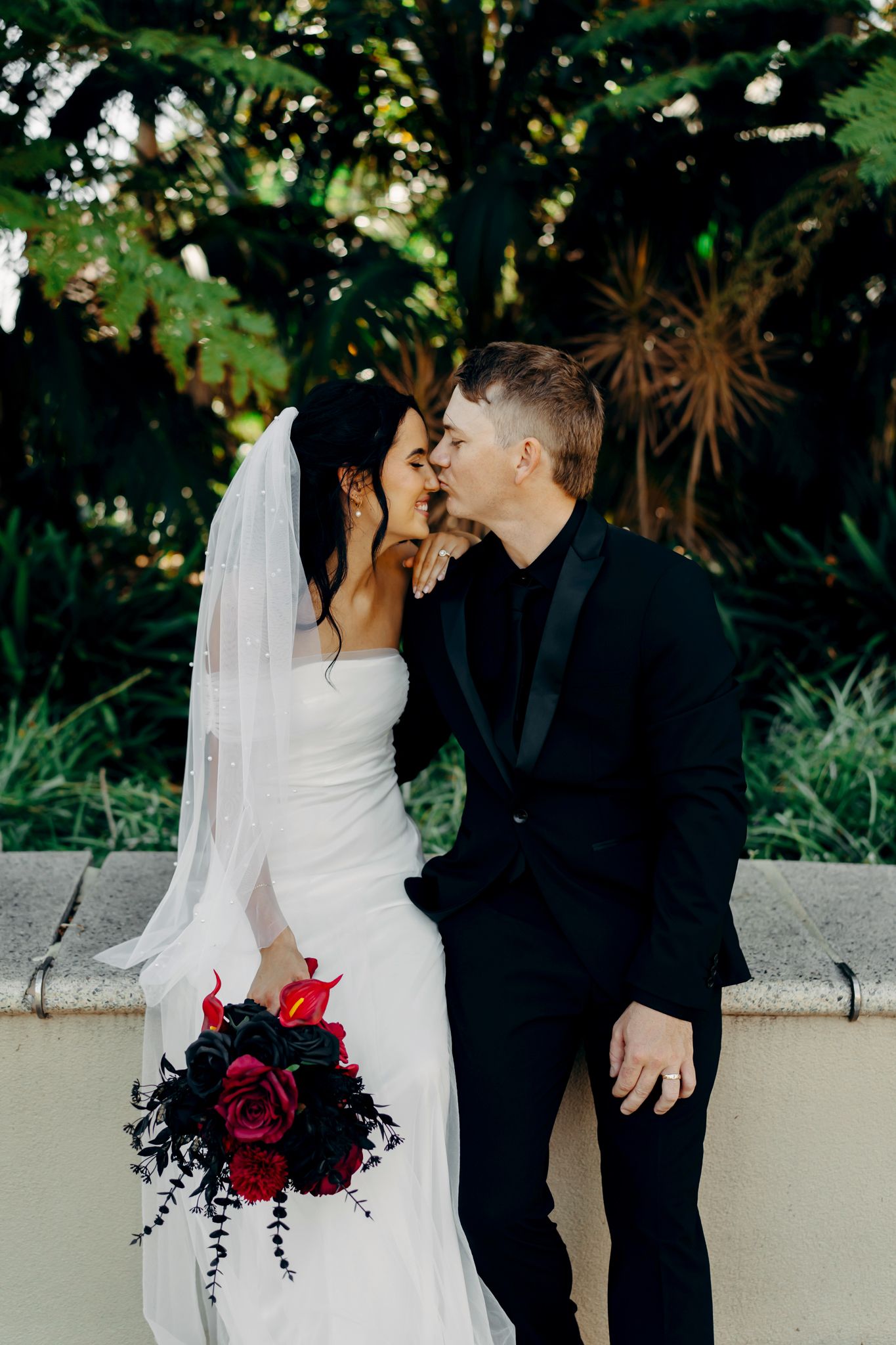 Bride and groom share a kiss outdoors, bride holding red rose bouquet in white gown with veil, groom in black suit.