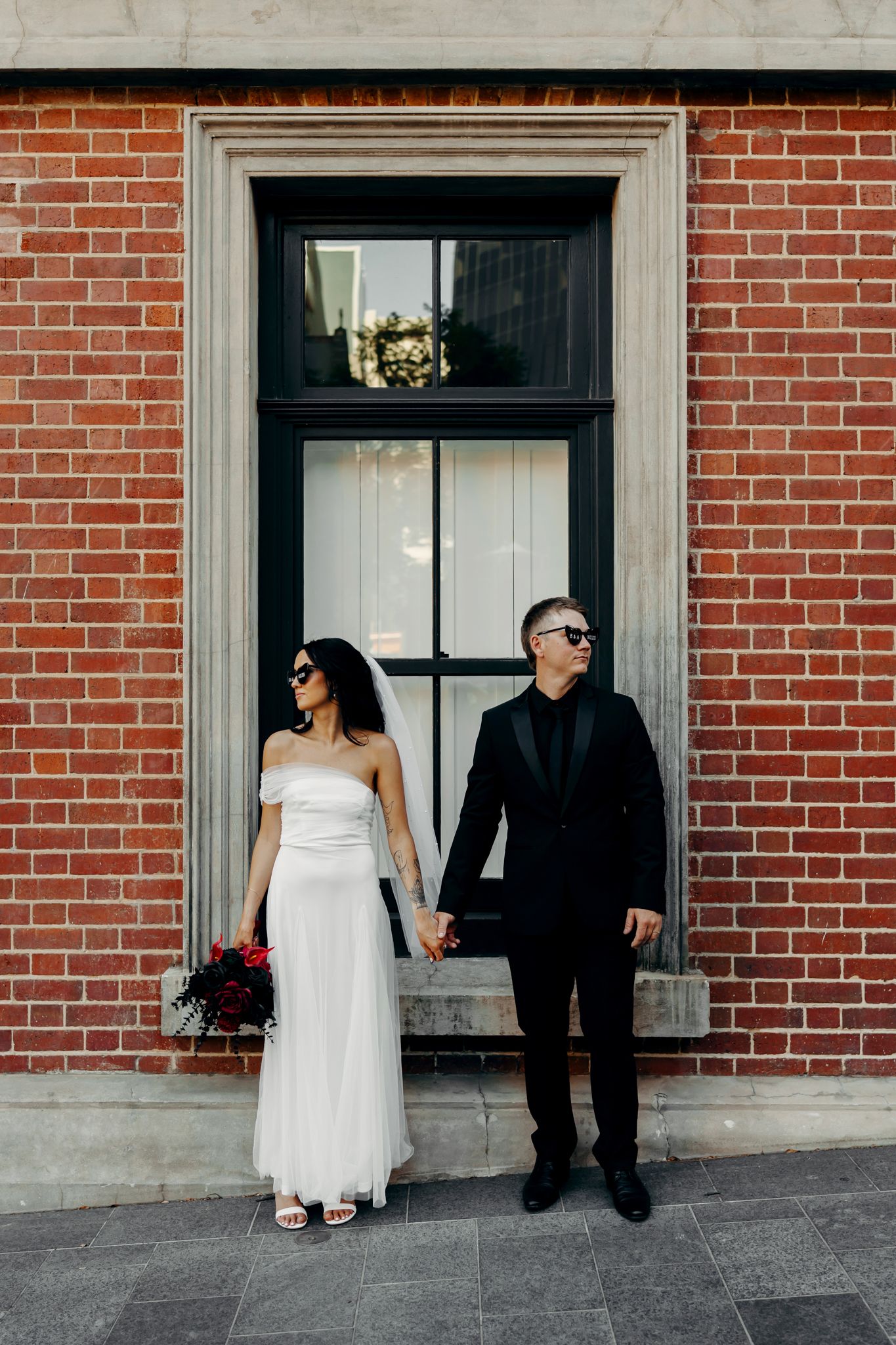Bride in white gown and groom in black suit holding hands before a red brick wall with sunglasses on.