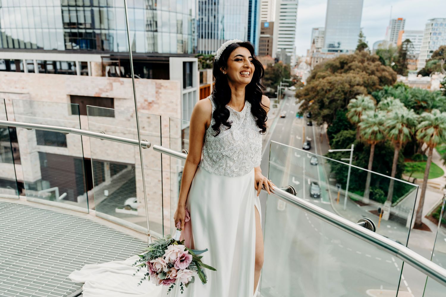 Elegant bride in a white beaded gown with thigh slit poses with bouquet on modern urban balcony.