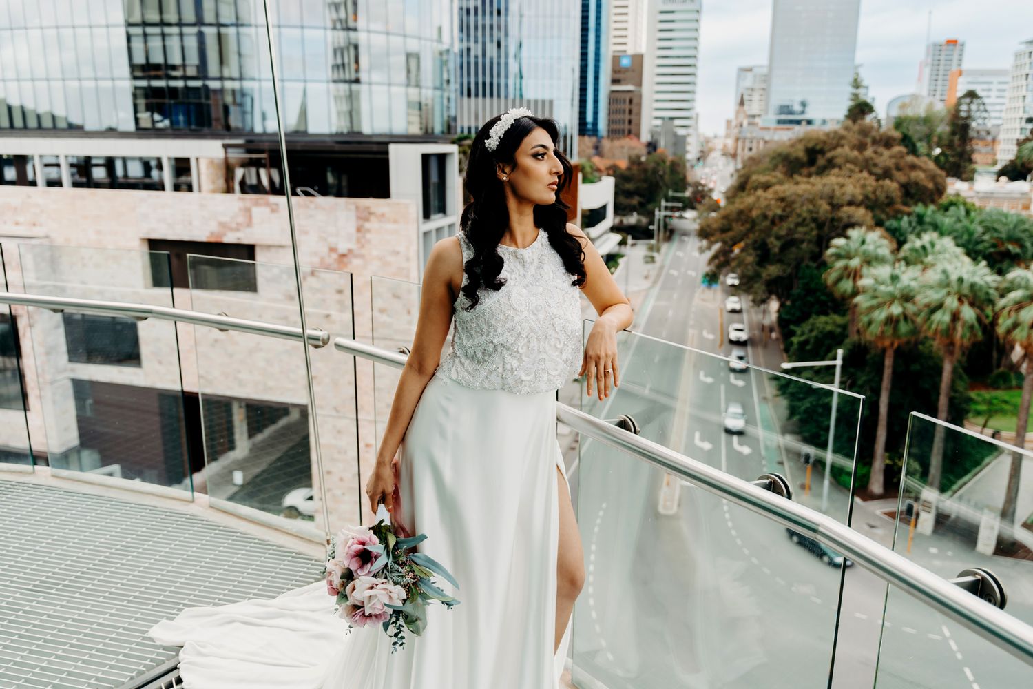 Bride holding pink bouquet stands on balcony overlooking city skyline in flowing white dress.
