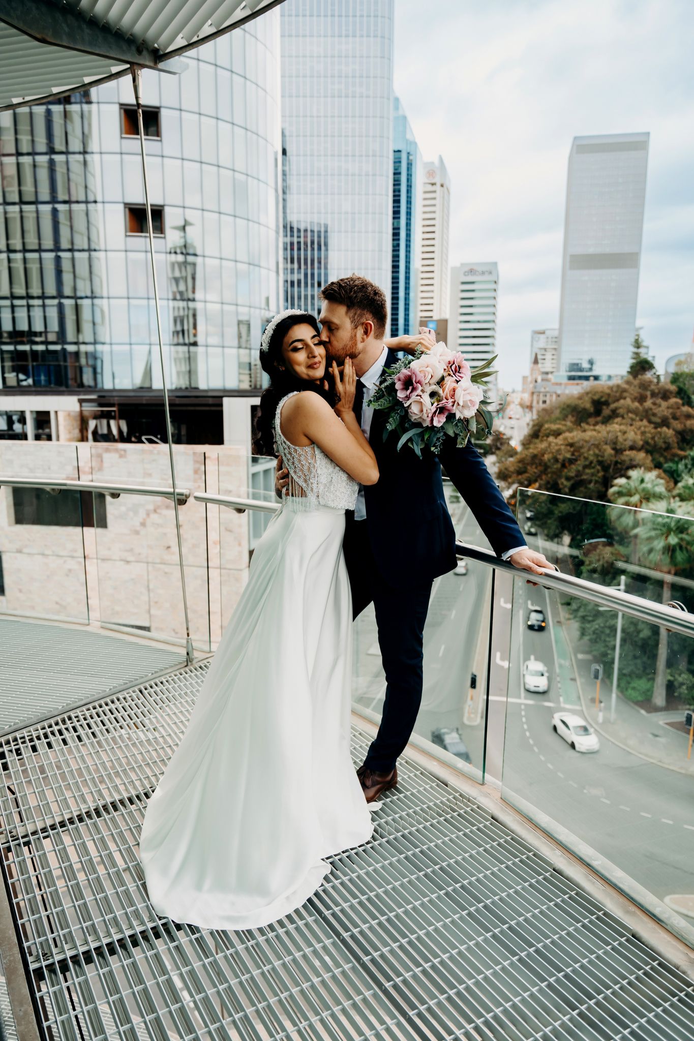 Romantic wedding couple embraces on modern architectural balcony with city views.