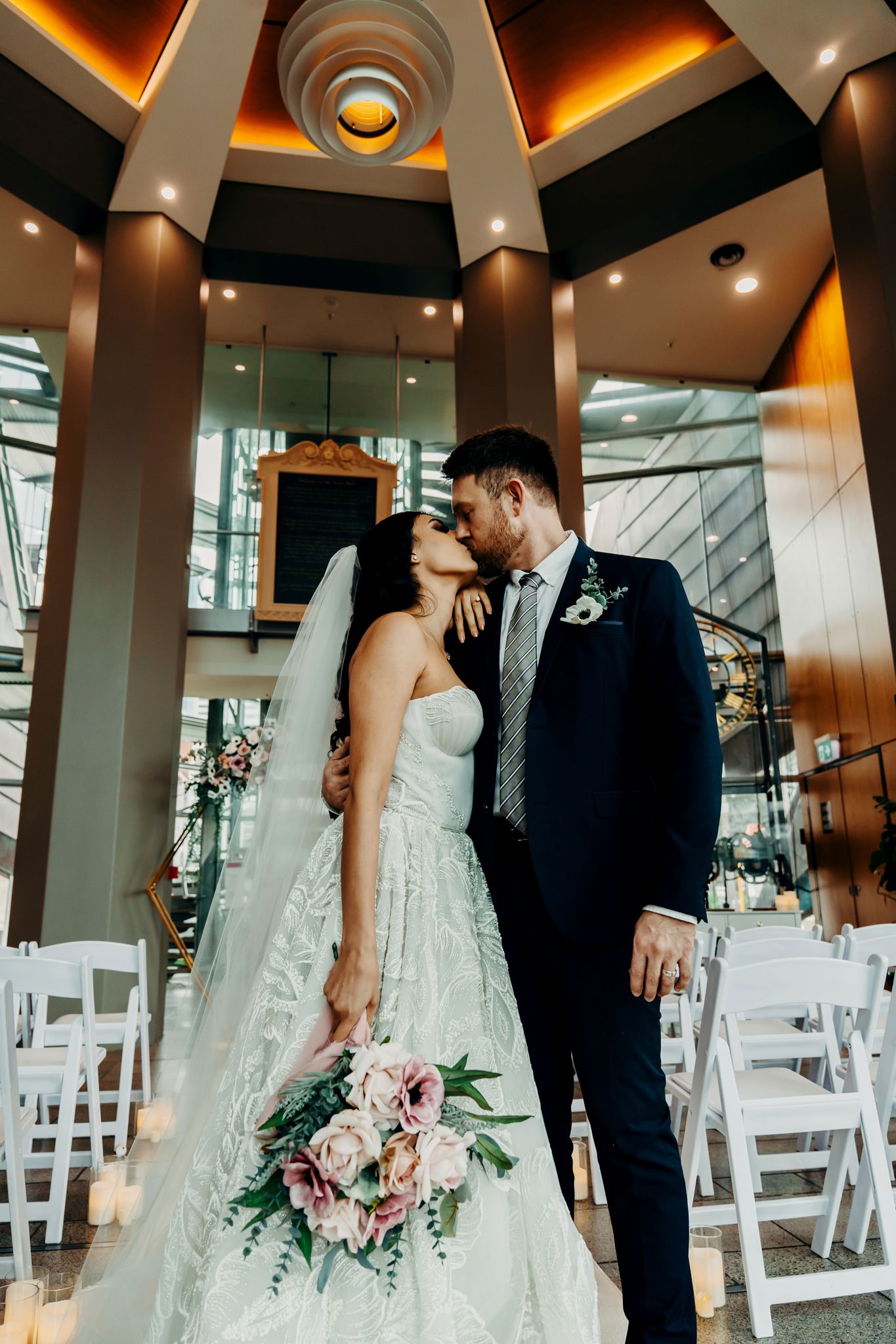 Romantic wedding portrait in modern venue with amber lighting and white ceremony chairs.