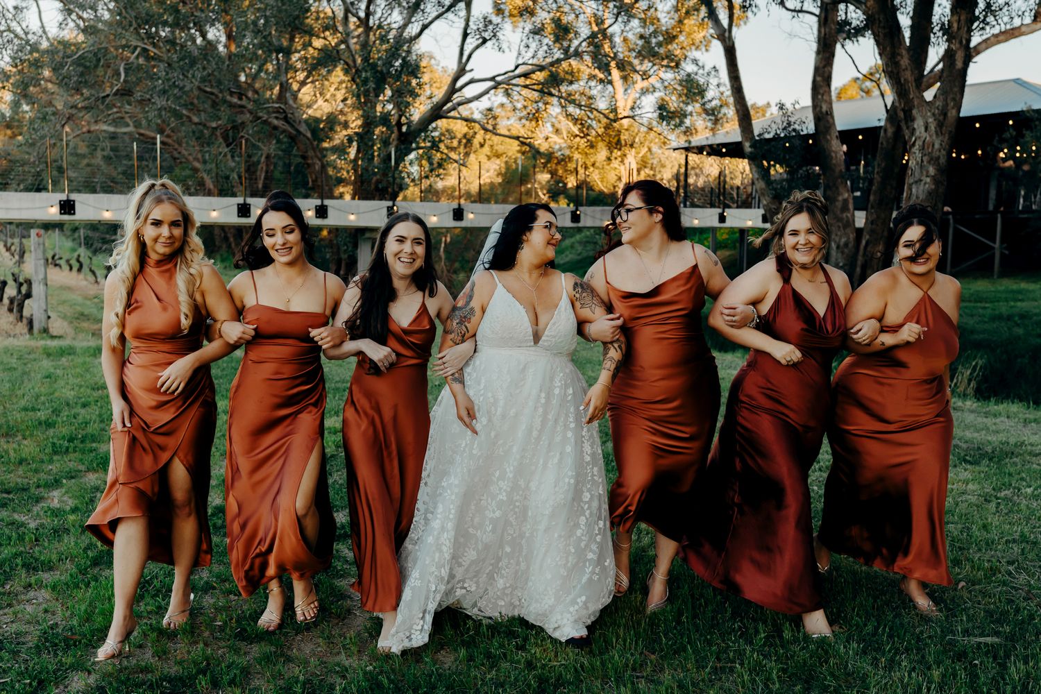Wedding party in matching rust-colored bridesmaid dresses poses with bride in white lace gown on grassy lawn at sunset.
