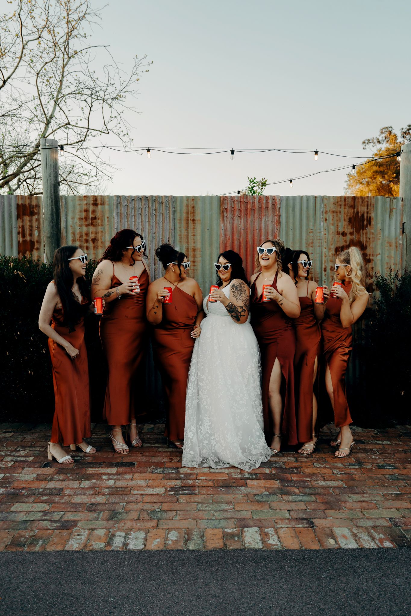 Wedding party in burgundy dresses poses playfully against industrial wall at sunset.