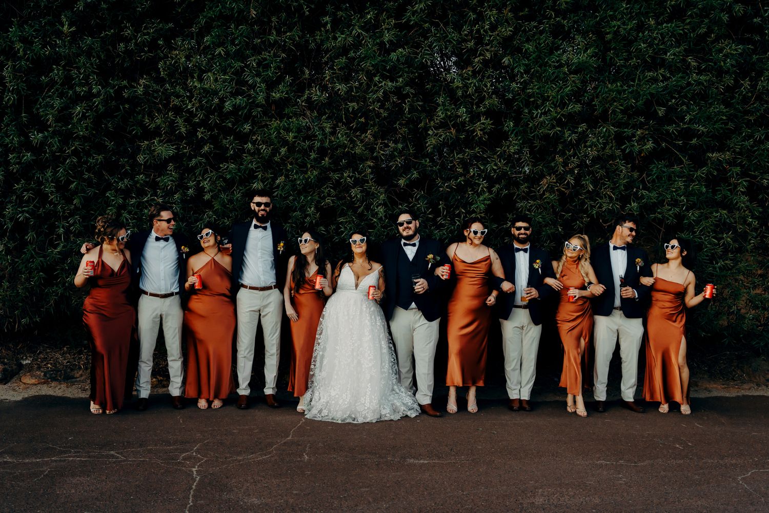 Wedding party in burnt orange and gray attire poses together in front of lush green hedges.
