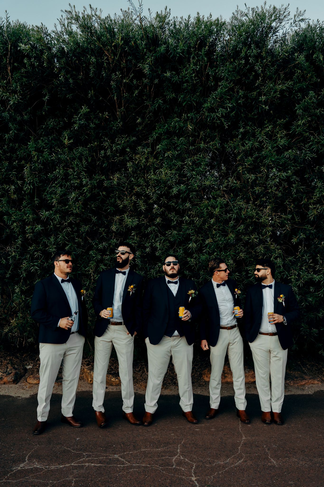 Groomsmen in matching navy jackets and light gray pants pose casually against a dark hedge backdrop.