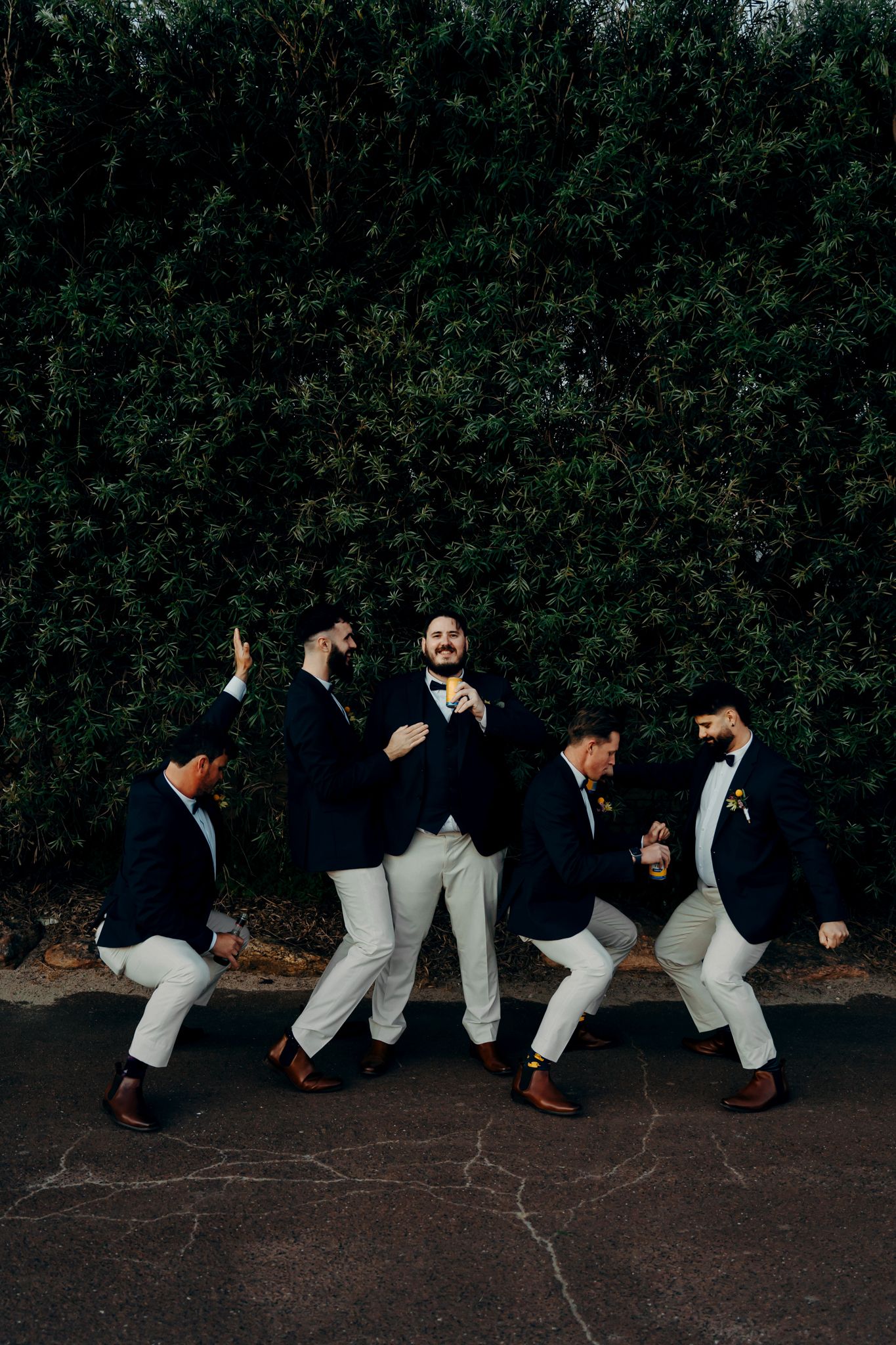 A group in matching navy blazers and white pants strike playful poses against a dark green hedge backdrop.