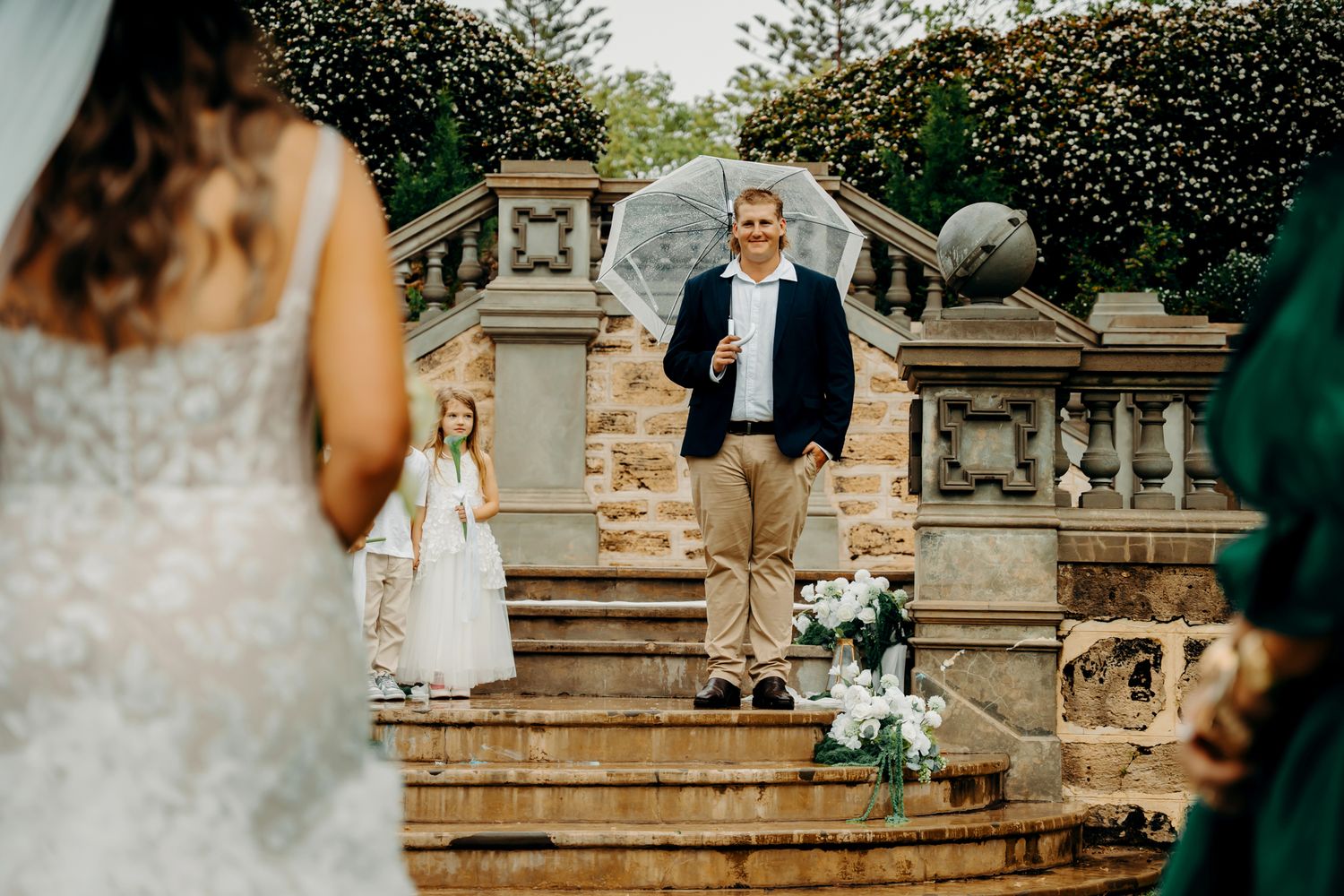 Elegant wedding party poses on ornate stone steps surrounded by lush greenery and decorative architecture.