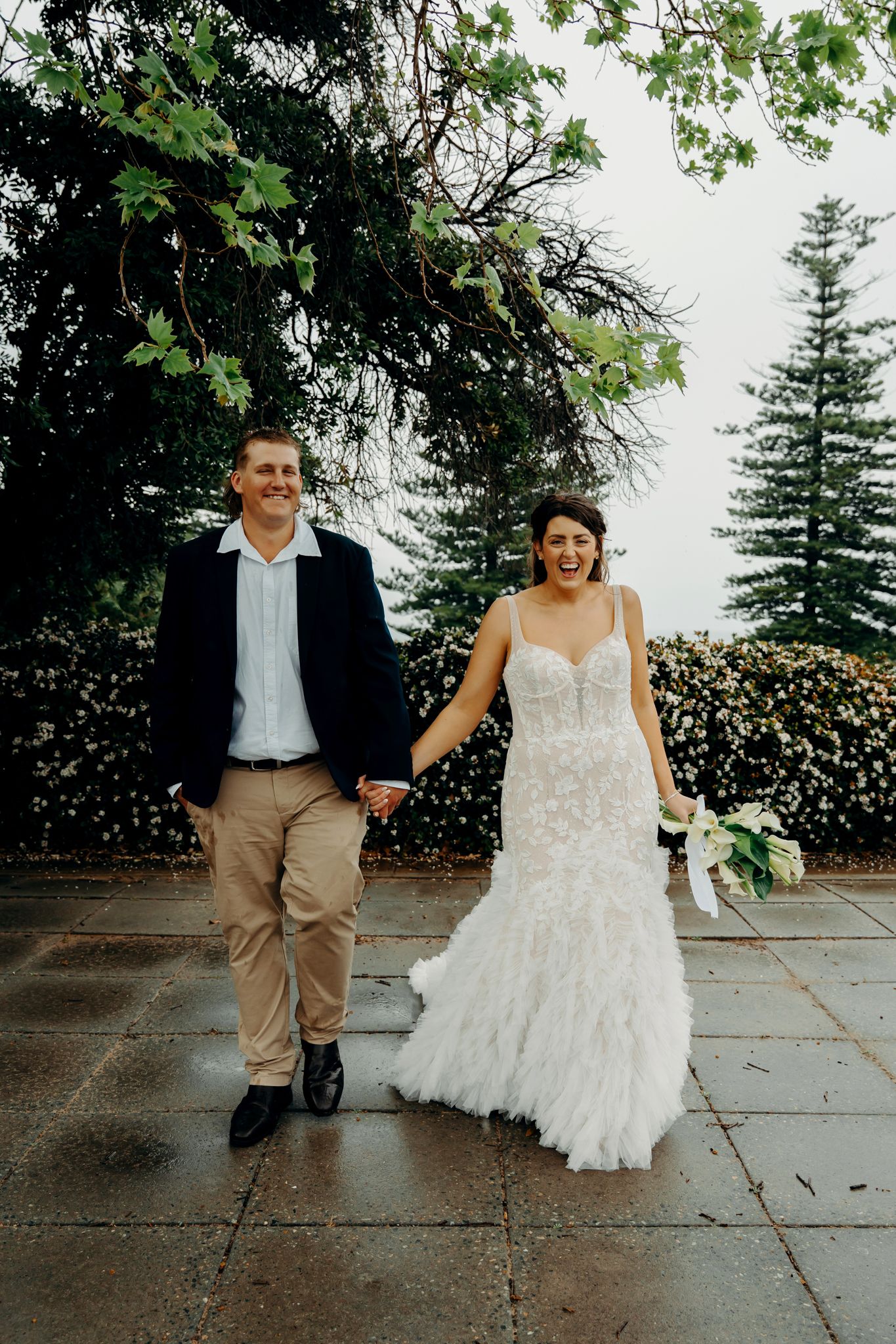 Wedding couple walking together on a stone path surrounded by trees and greenery.