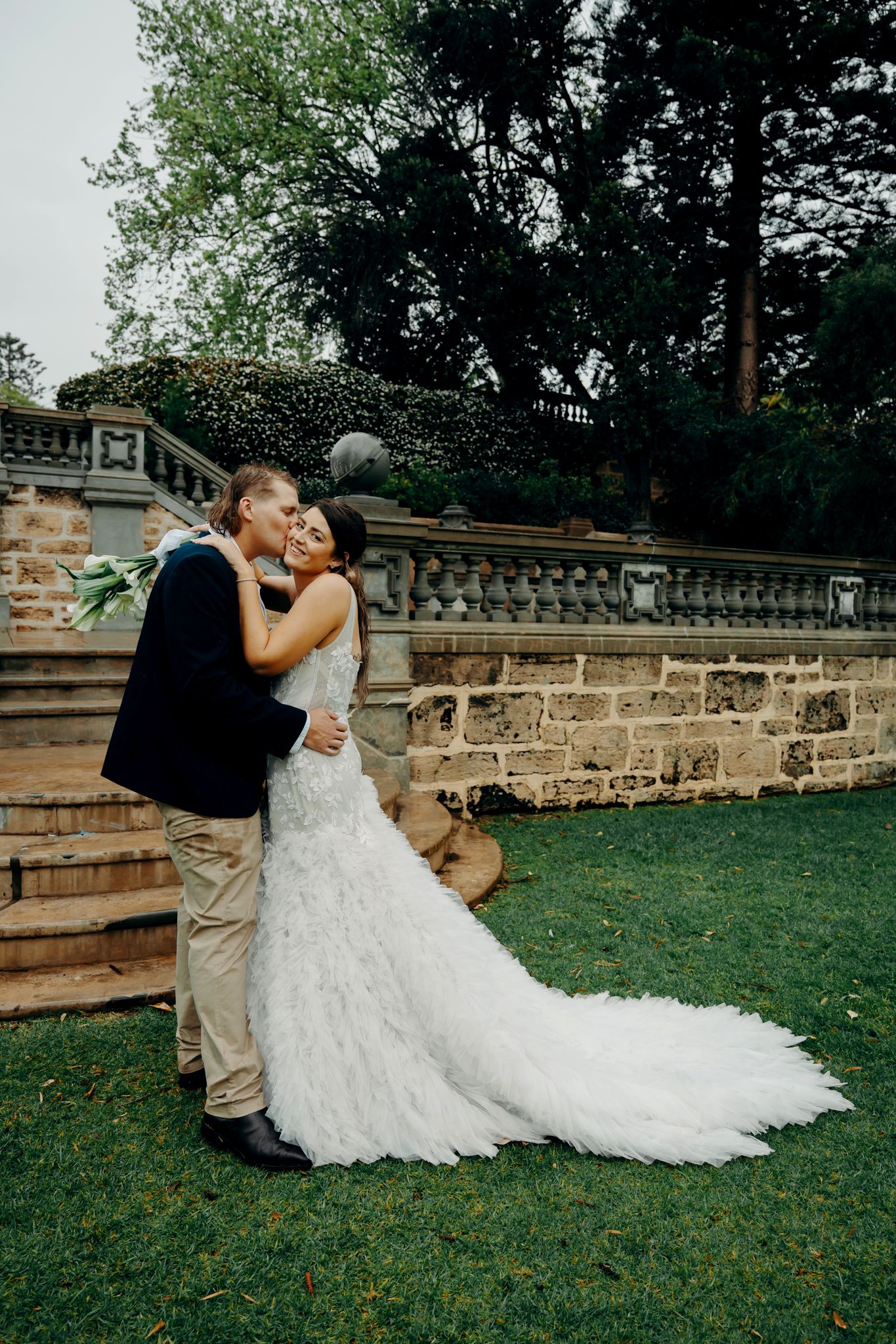 A romantic wedding couple shares intimate moments on a grassy lawn near a stone wall during their outdoor photo session.