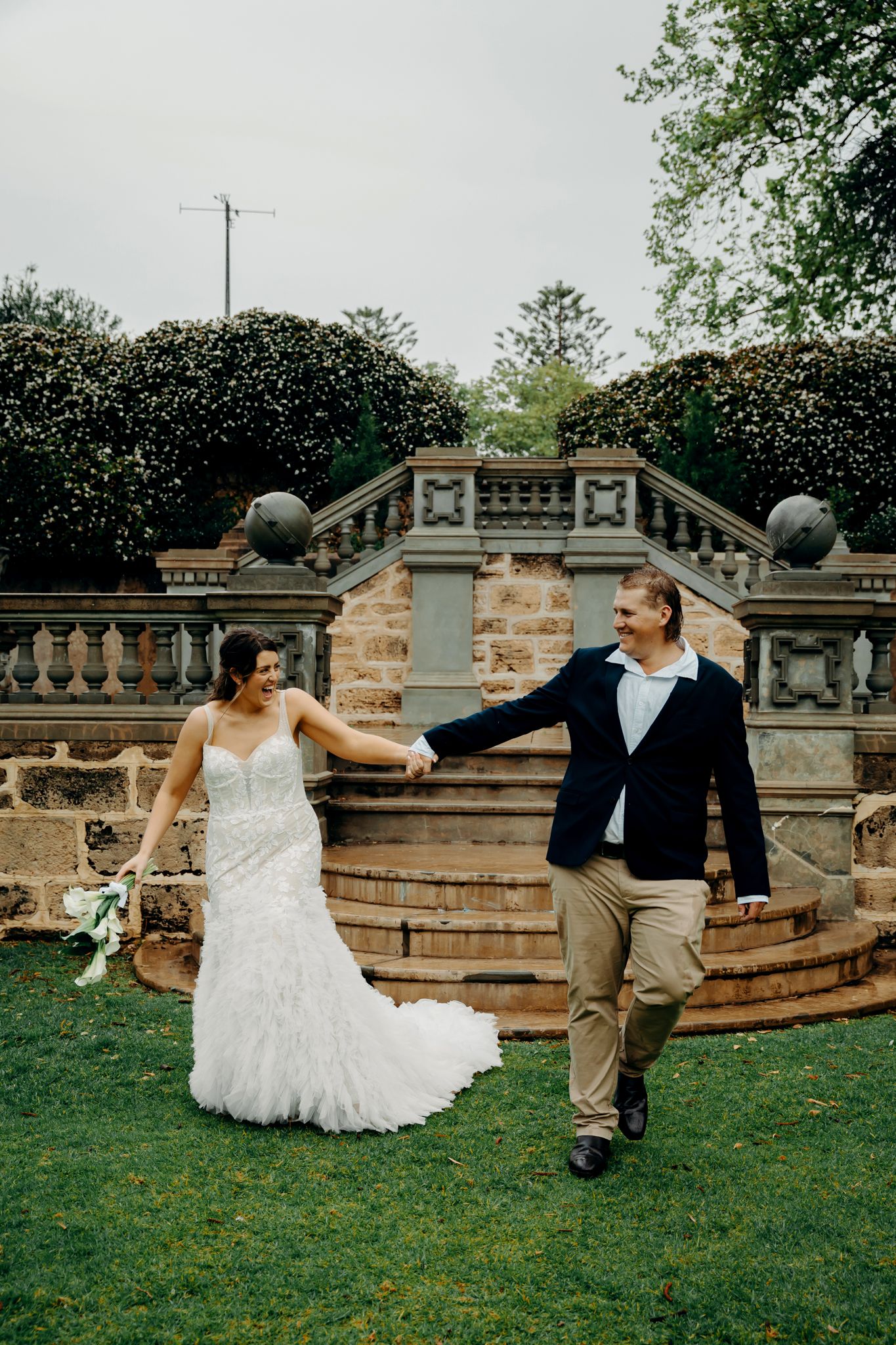 A playful wedding couple dances and twirls on the lawn beside a decorative stone wall during their celebration.
