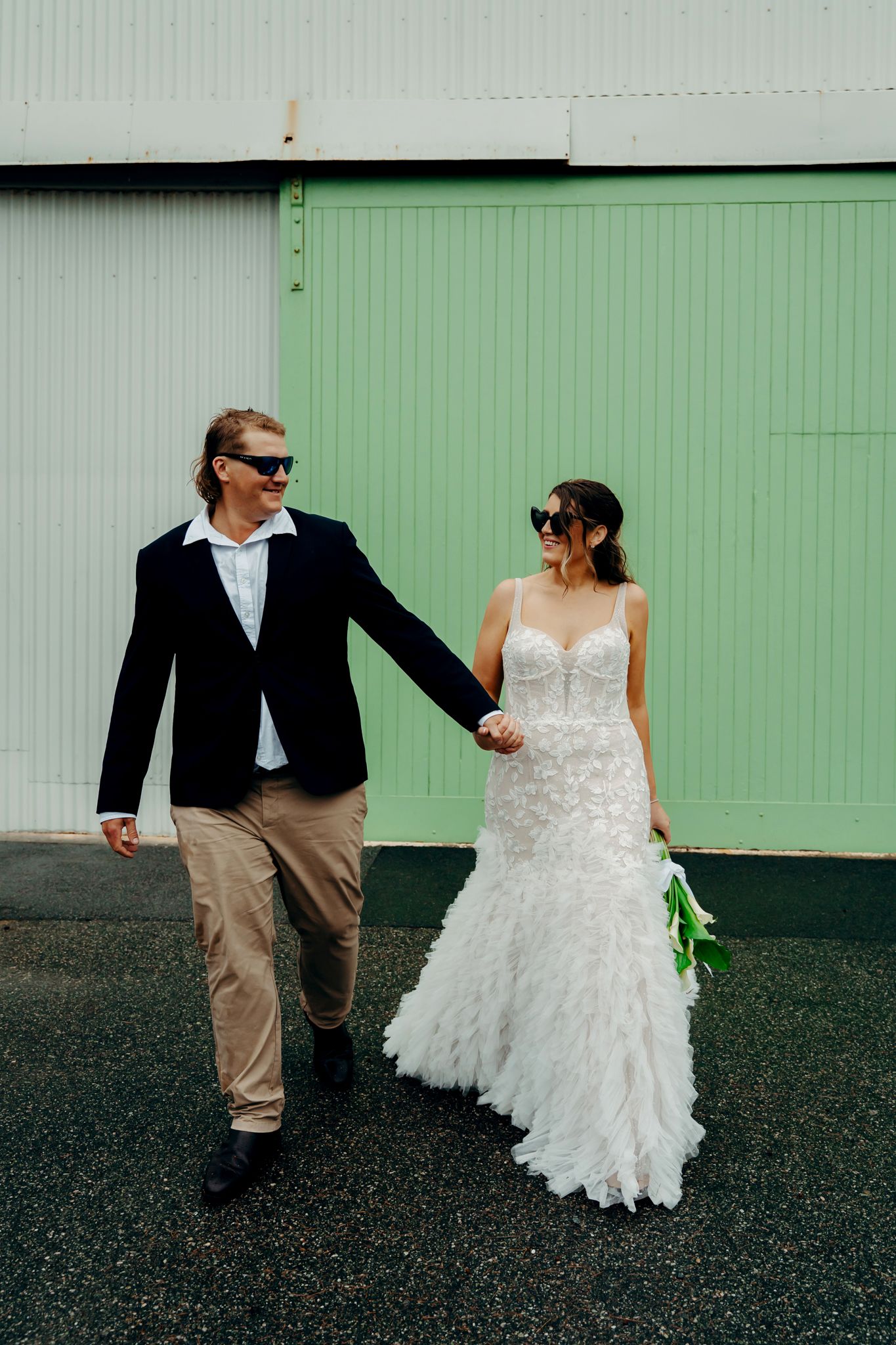 A couple in wedding attire walk hand in hand against a mint green wall, with the bride in a lace gown and groom in sunglasses.