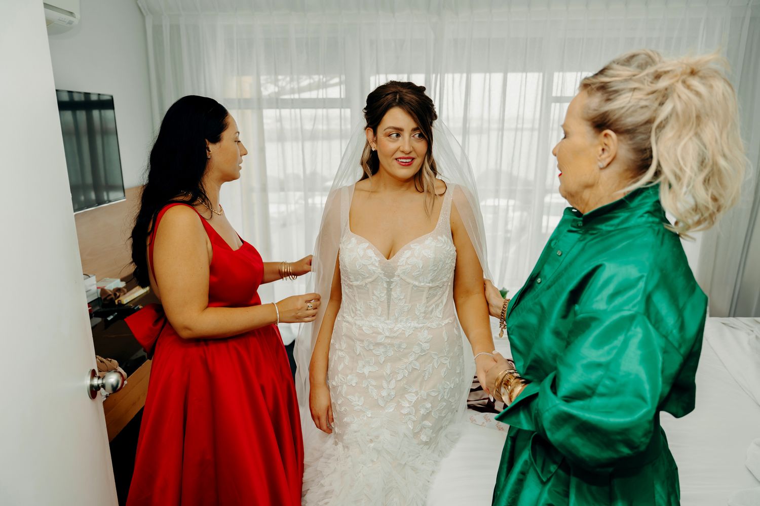 A bride in a white lace gown stands with two guests wearing red and emerald dresses during wedding preparations.