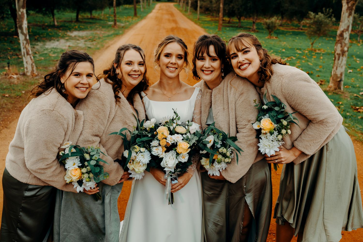 A joyful wedding party in neutral beige dresses poses together outdoors while holding floral bouquets.