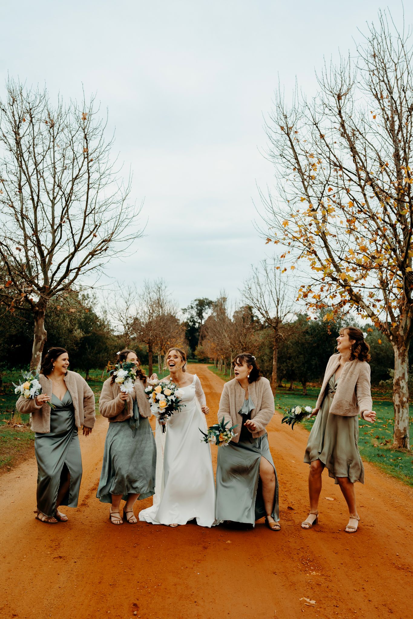 Wedding party walks and laughs together on a rustic dirt path during autumn with grey and tan attire.