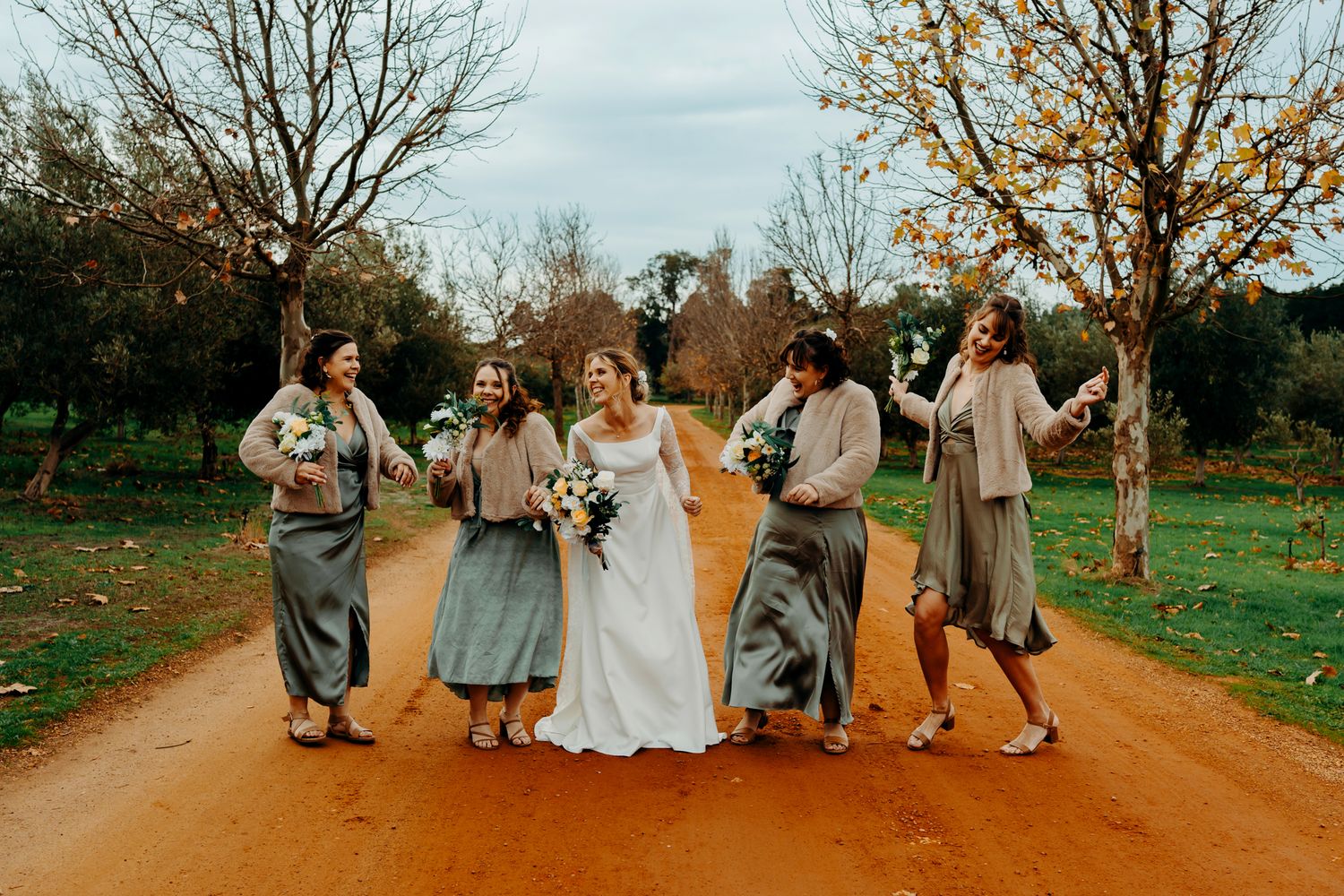 A wedding party joyfully walks and dances together on a rustic orange dirt road surrounded by autumn trees.