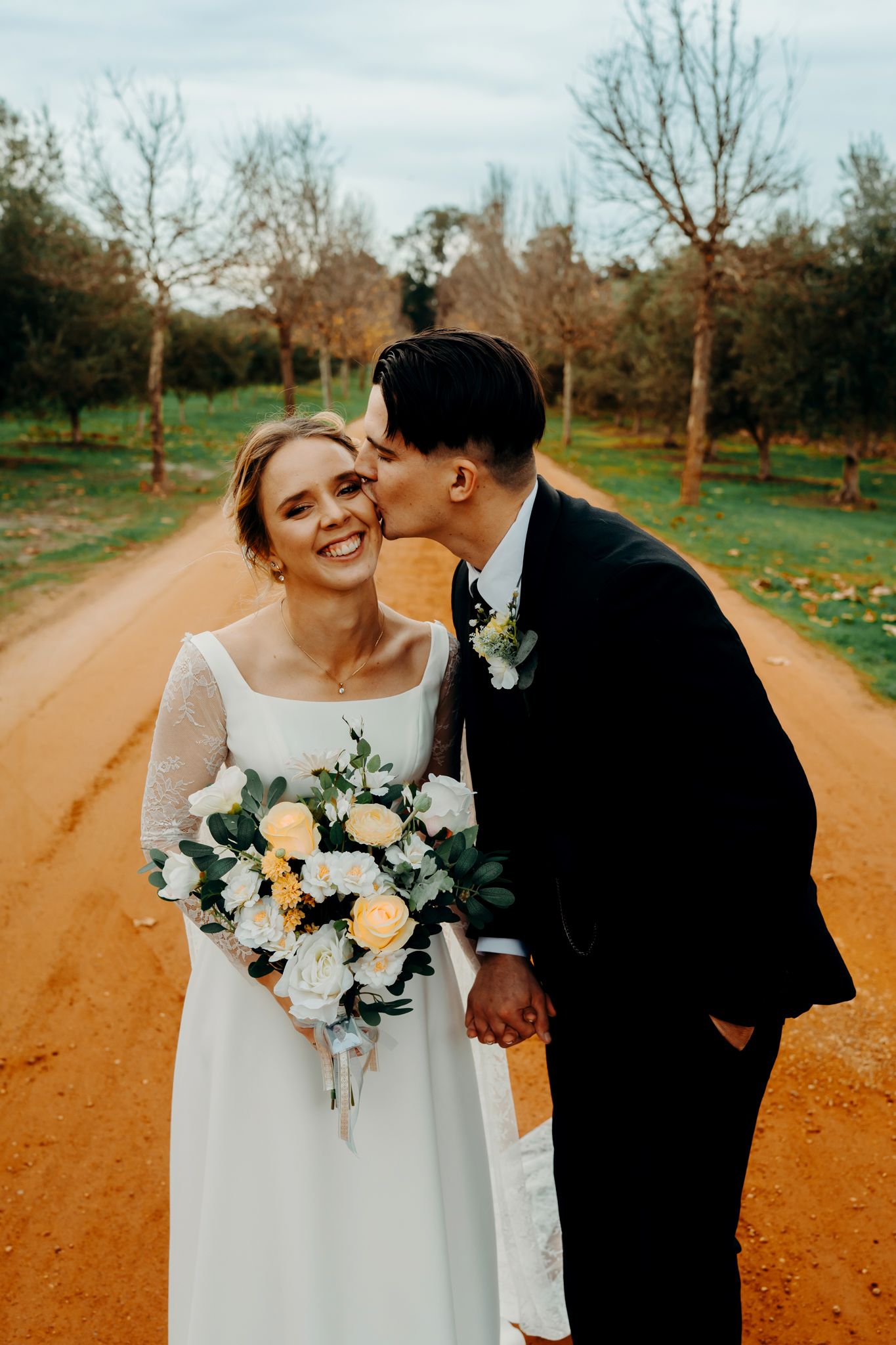 A rustic orange dirt road winds through a park with trees lining both sides while a couple celebrates their wedding day.
