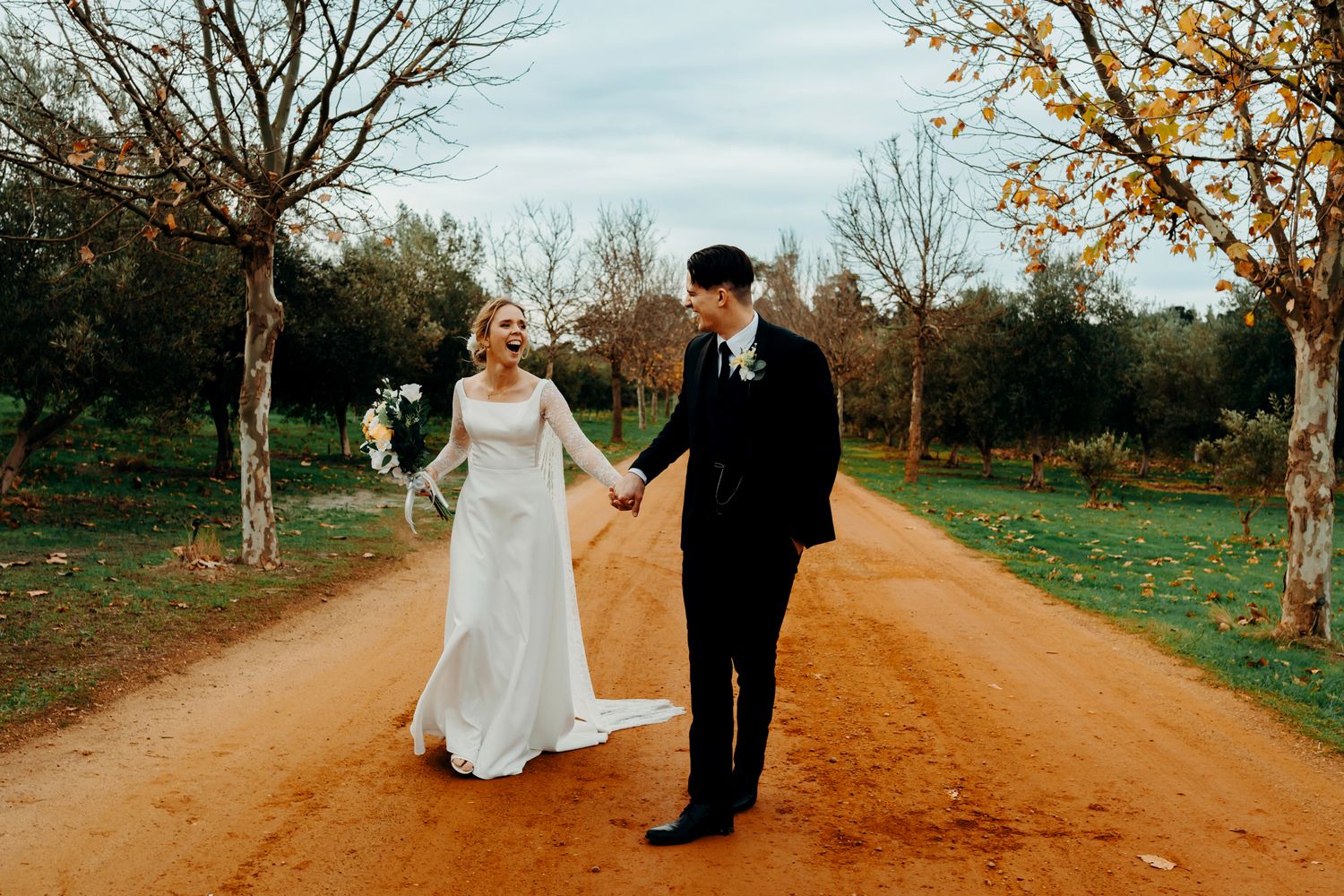 Couple in formal wedding attire holding hands while walking down a rustic dirt path lined with autumn trees.