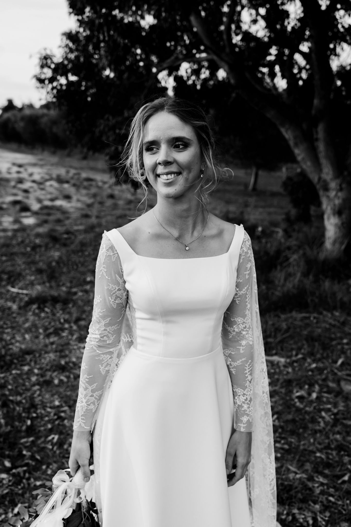 A bride in a long-sleeved white wedding dress with lace details stands outdoors against a dark background.