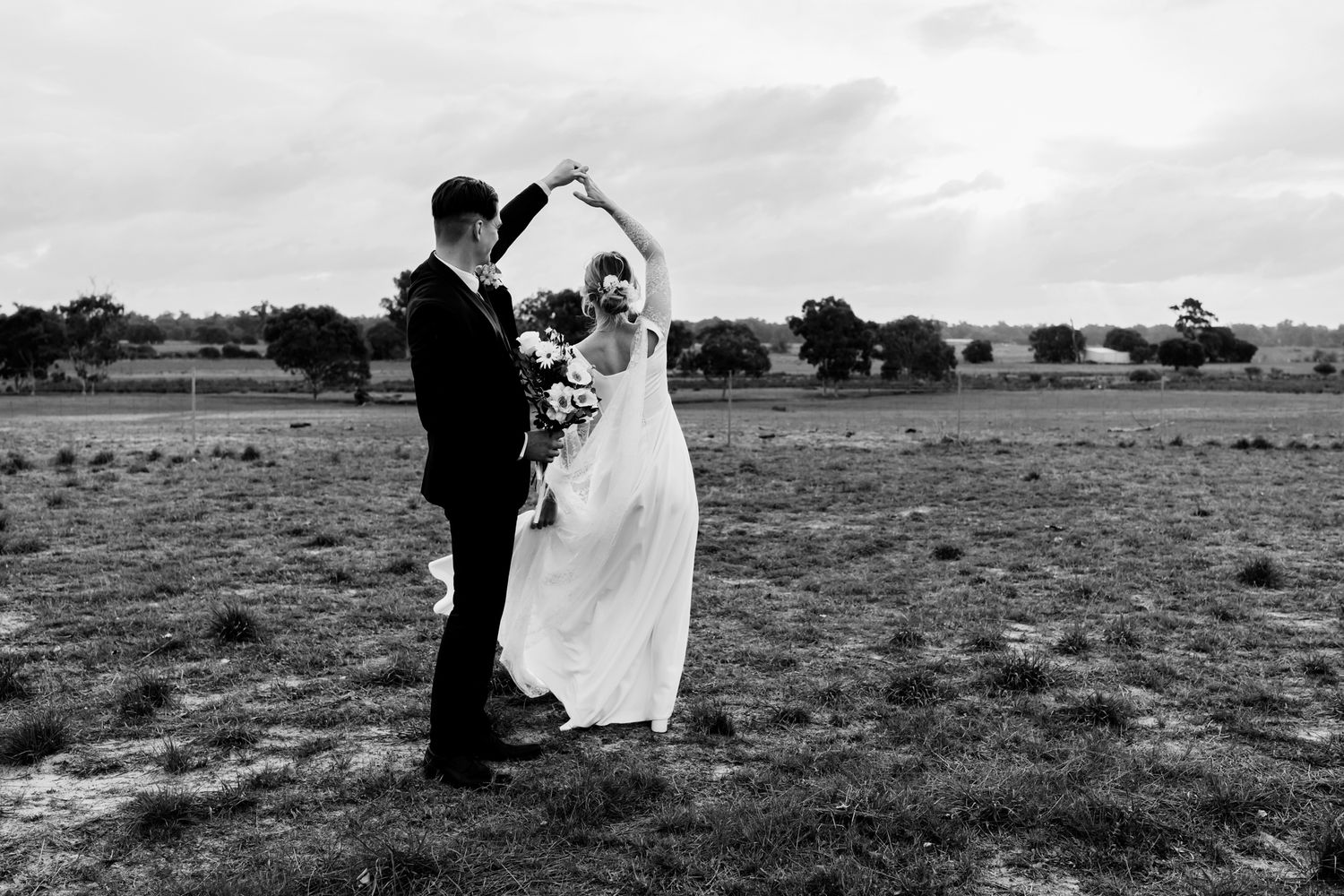 A bride and groom dance together in an open field on their wedding day, captured in black and white photography.