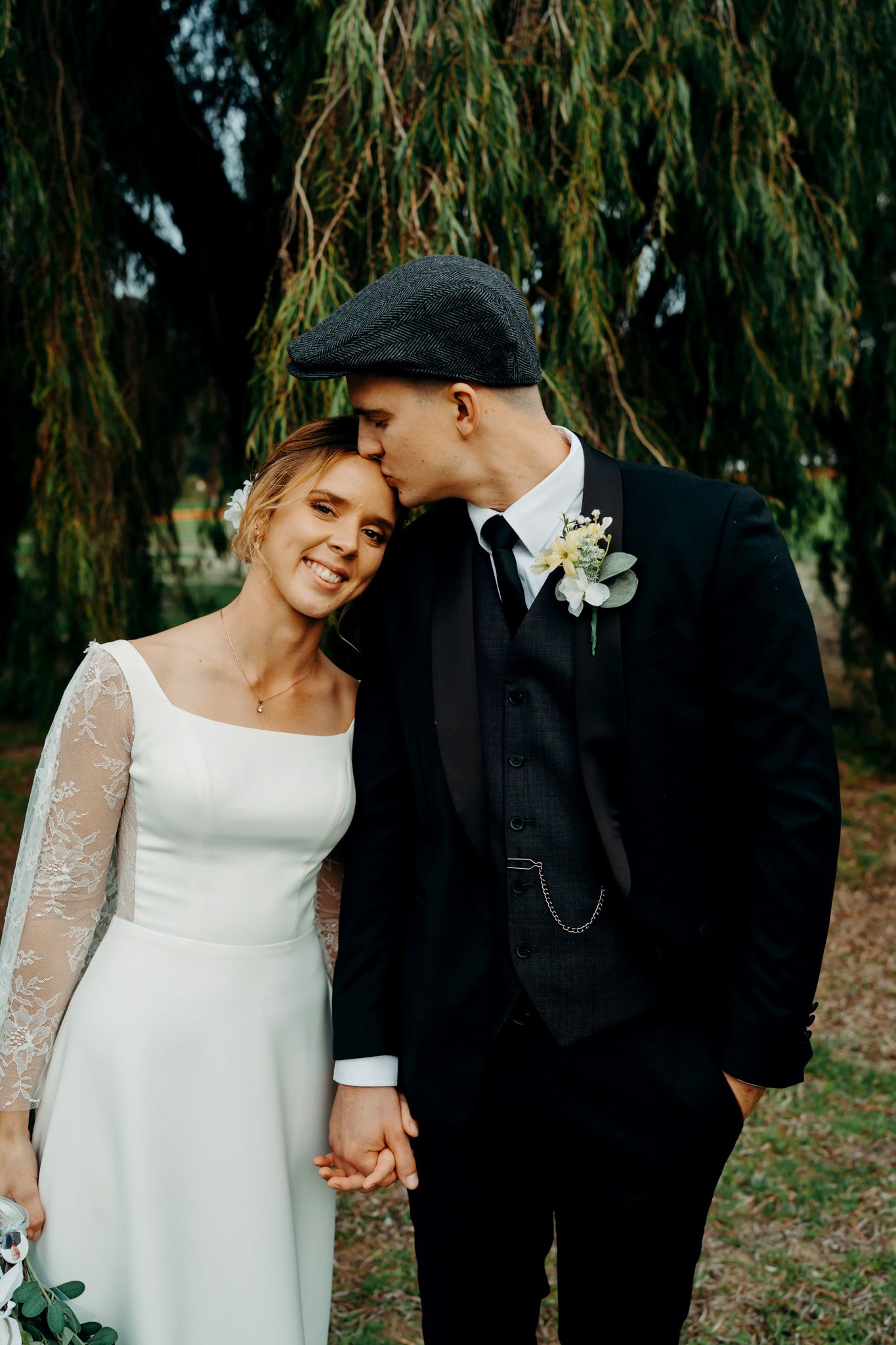 A romantic wedding portrait of a couple under a weeping willow tree, with the bride in white and groom in black attire.