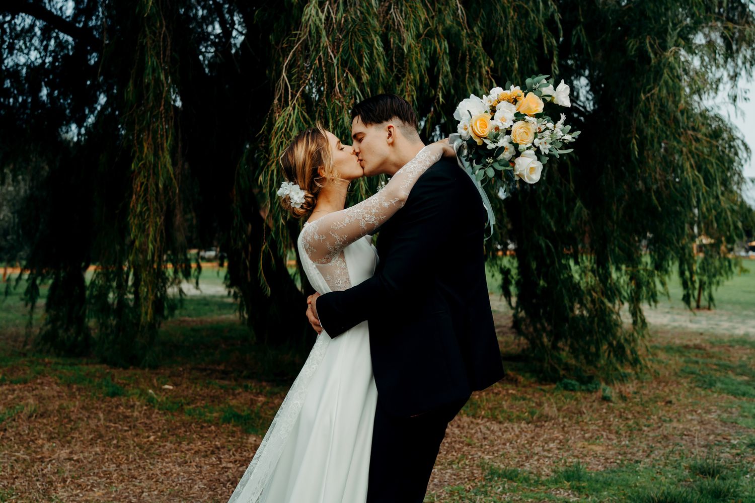 Two newlyweds share a romantic kiss under a weeping willow tree while the bride holds a white and yellow bouquet.