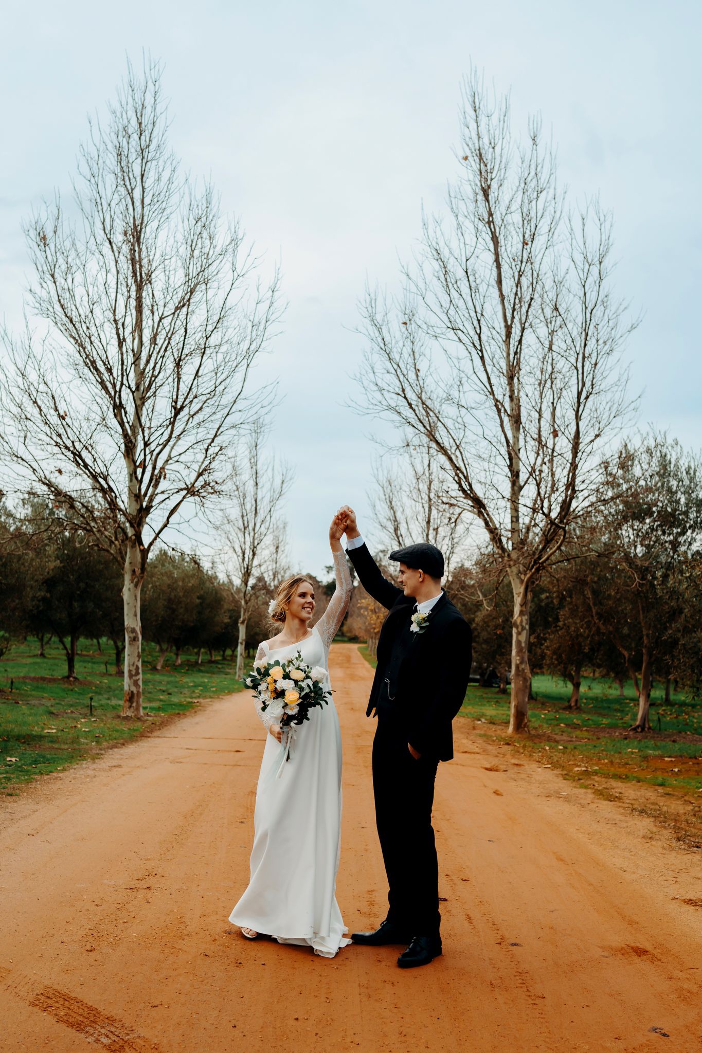 Romantic wedding couple dances along a scenic dirt path lined with bare trees on an overcast day.