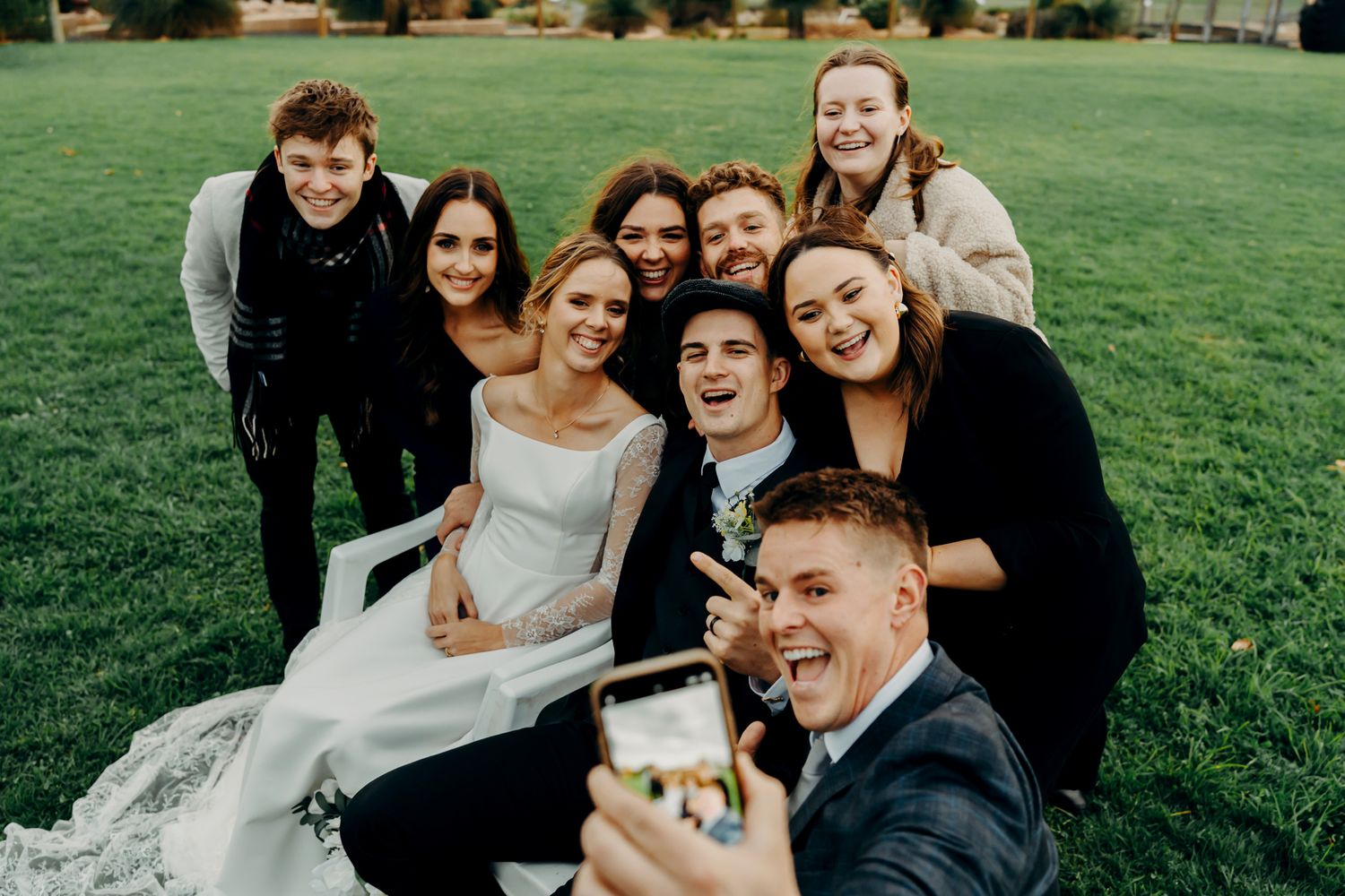 A group of friends posing together on green grass while taking a selfie at an outdoor wedding celebration.