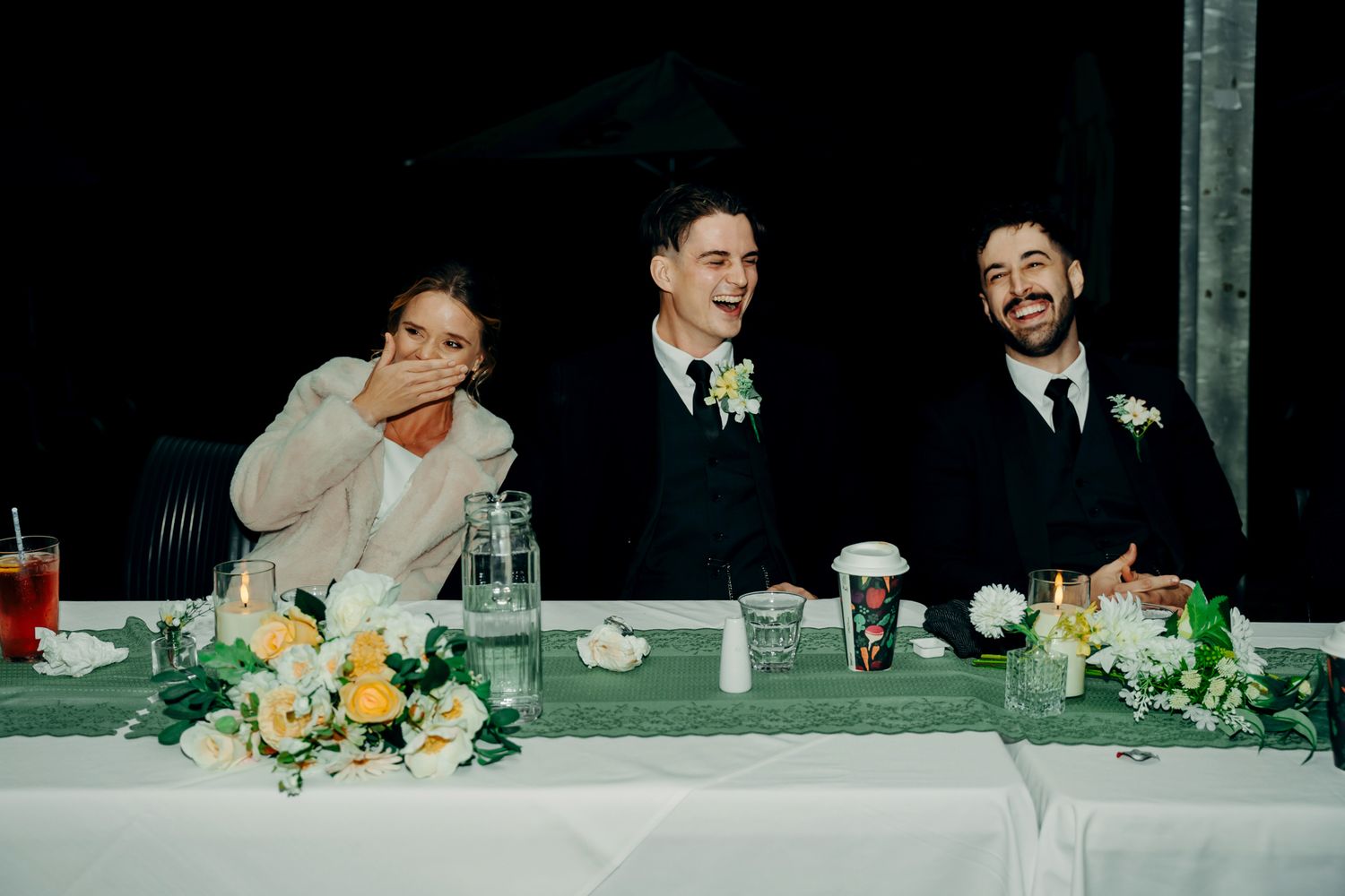 A candid moment at a wedding reception head table shows three people laughing together beside yellow floral centerpieces.