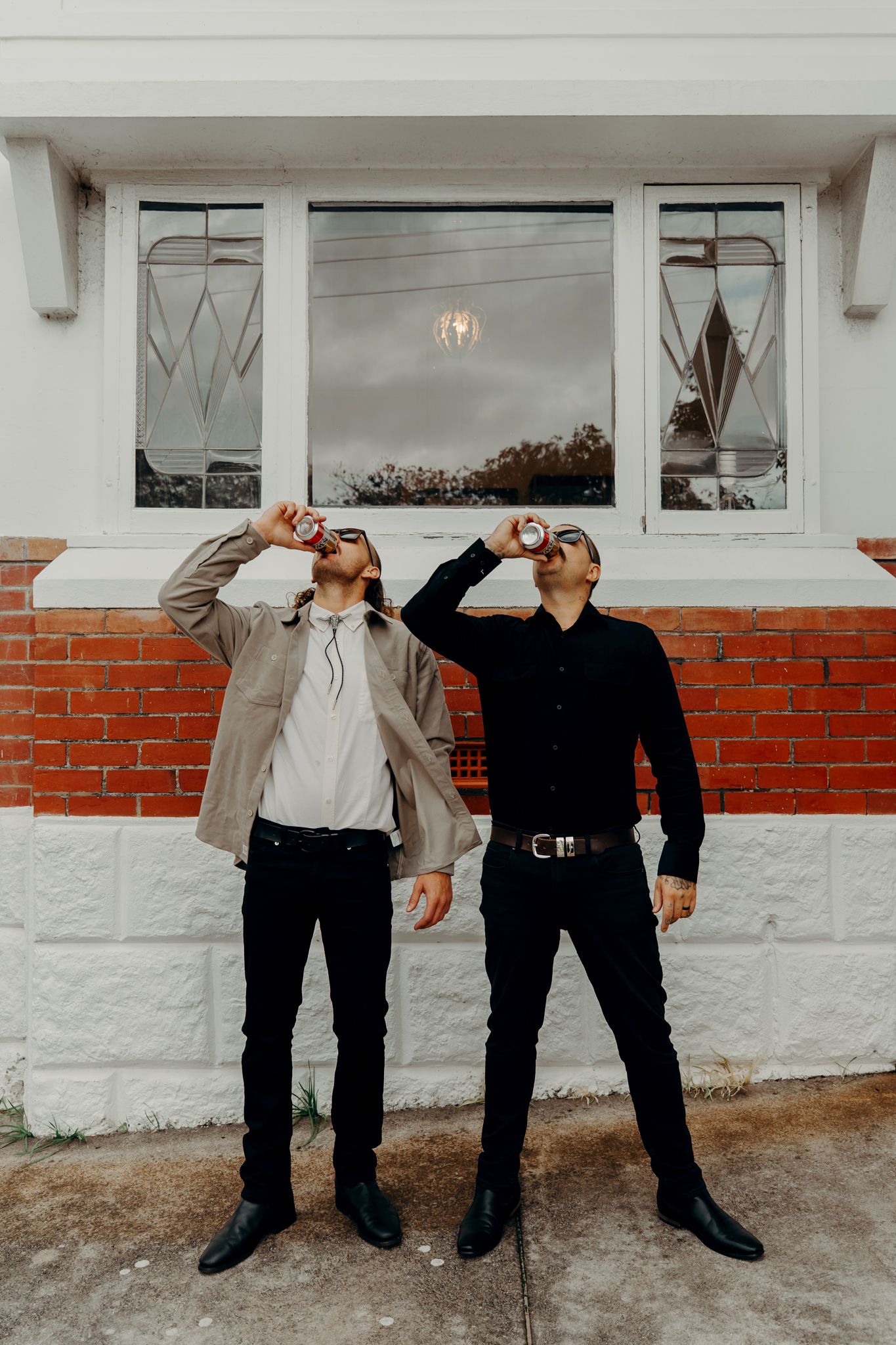 Two people in casual attire strike synchronized poses leaning against a white wall with a red brick accent and window.