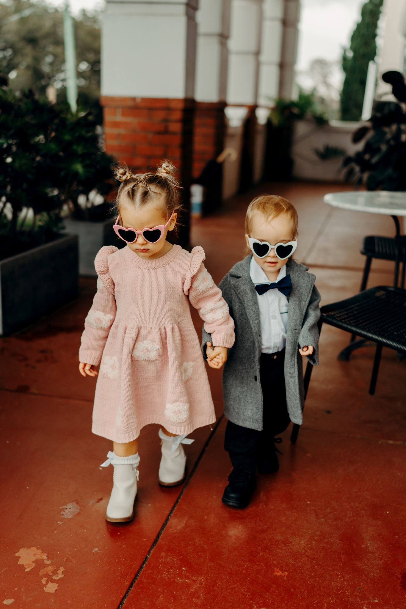 Two young children in dressy coats and sunglasses walk together holding hands on a red patio floor.
