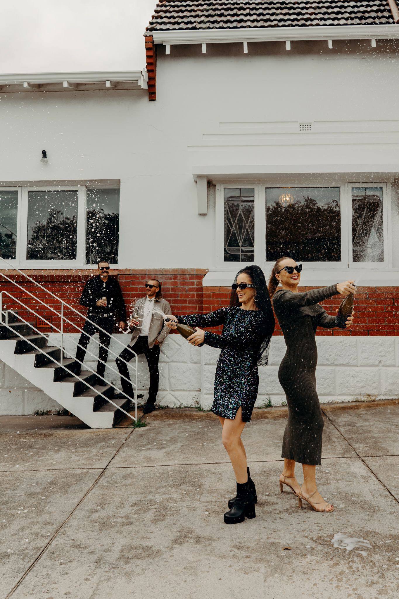 Group of friends in stylish dresses posing and dancing together outside a white house with brick accents on a sunny day.