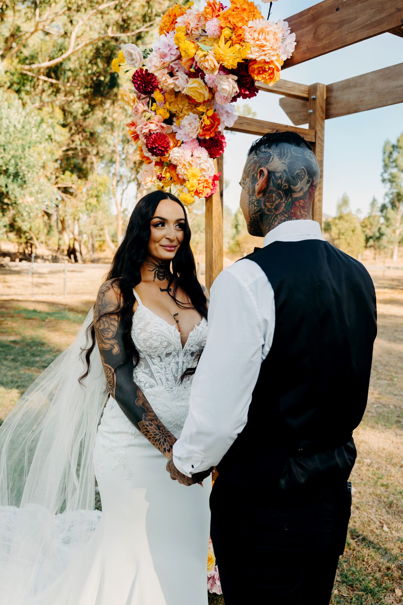 A couple exchanges vows under a wooden arch adorned with vibrant orange and pink flowers in an outdoor garden setting.