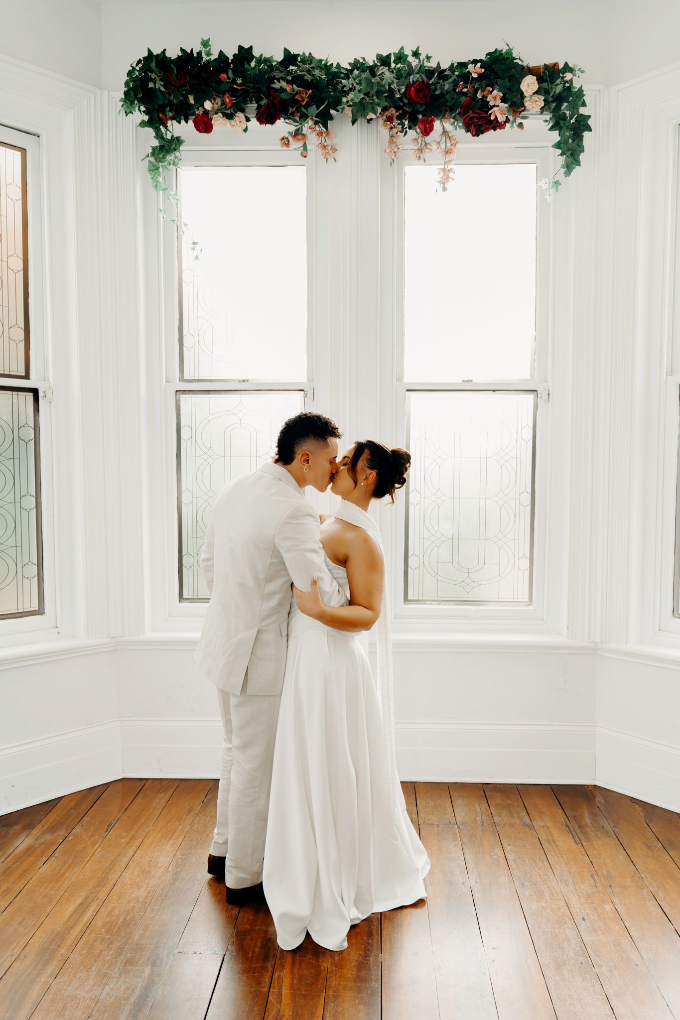 Bride and groom share a romantic kiss in a white room with hardwood floors and floral garland above bay windows.