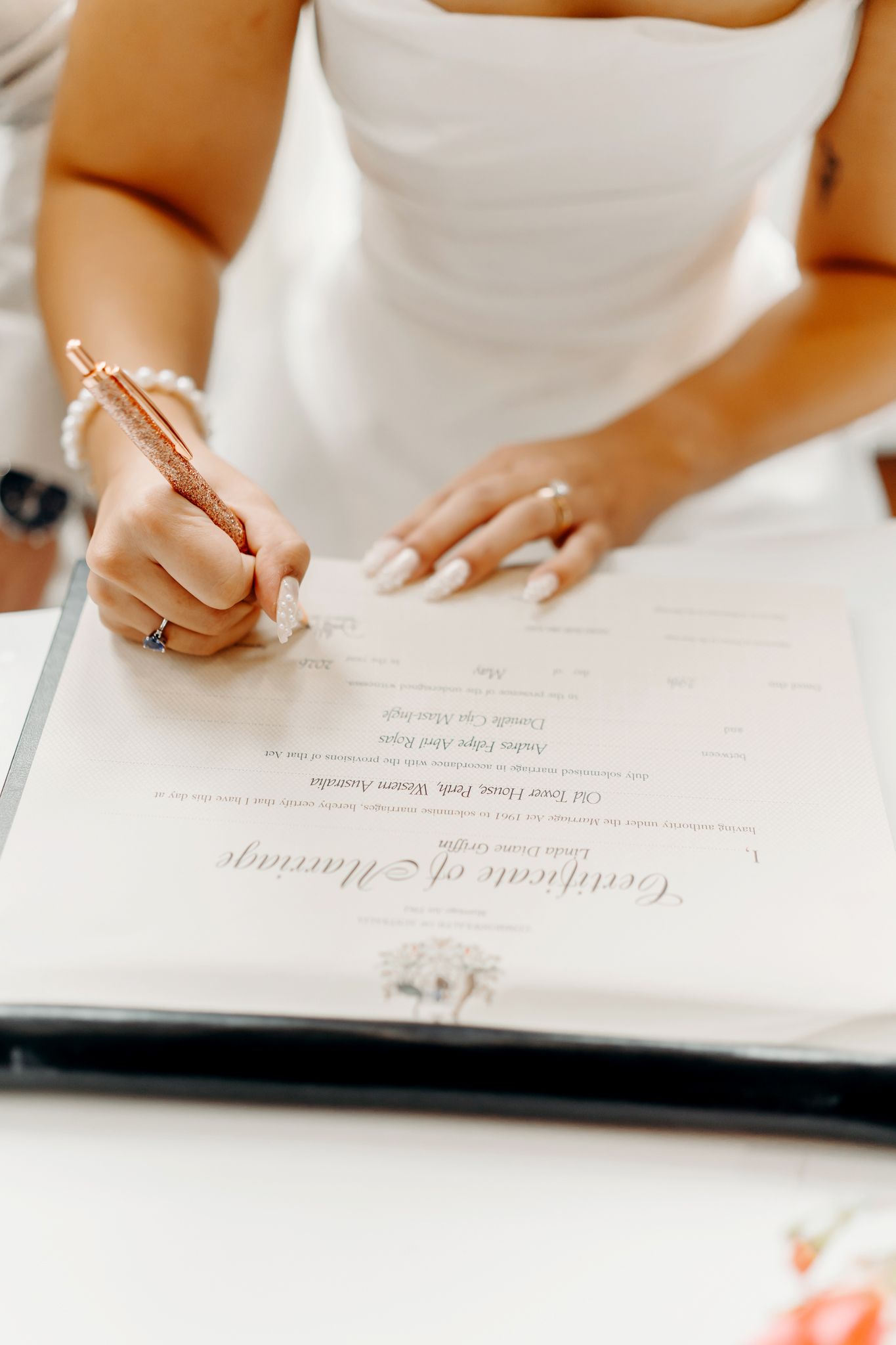Bride signing a marriage certificate with a rose gold pen, wearing a white wedding dress and pearl bracelet.