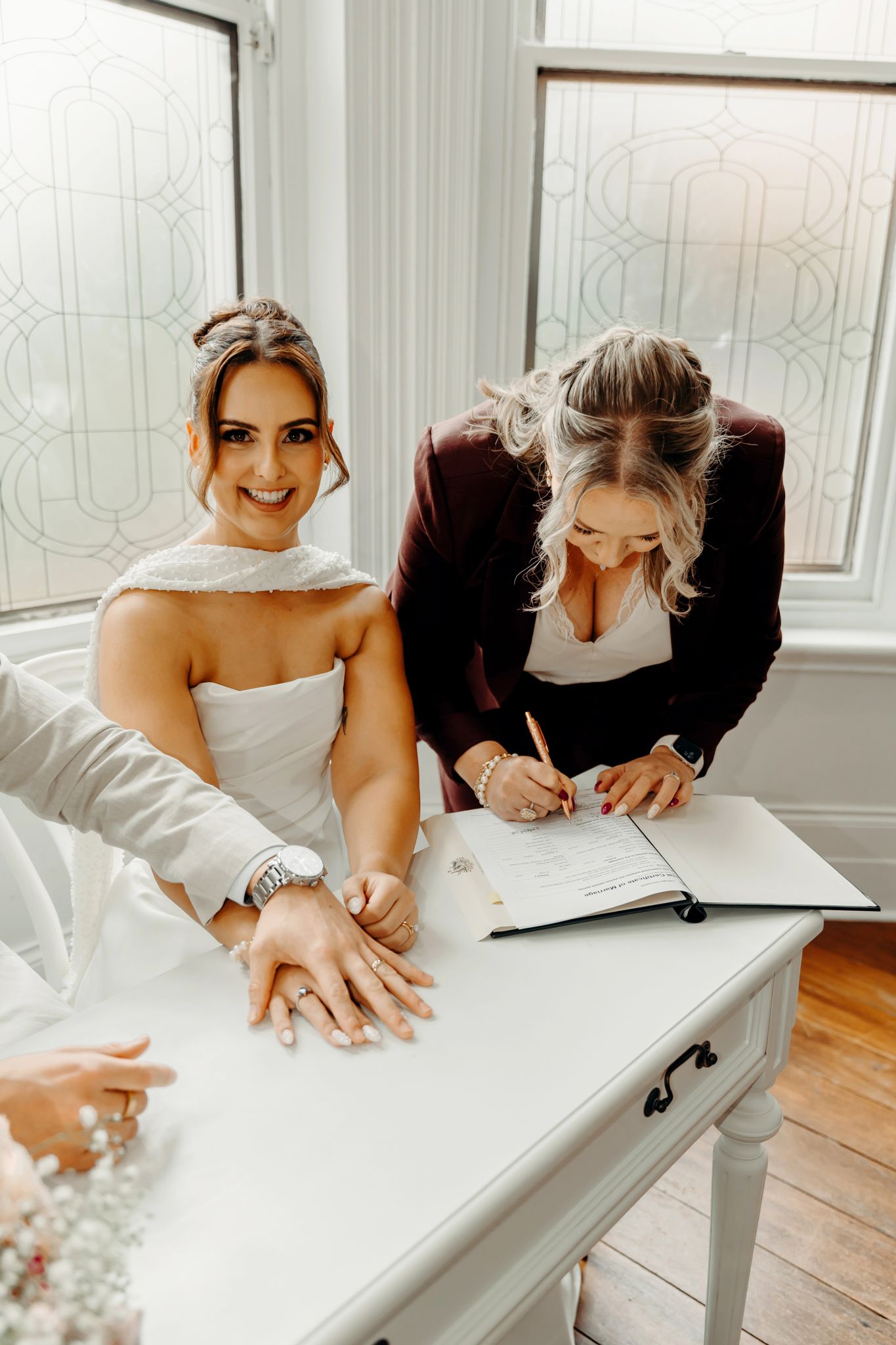 Bride smiling at camera while officiant signs wedding register at white table during indoor ceremony.