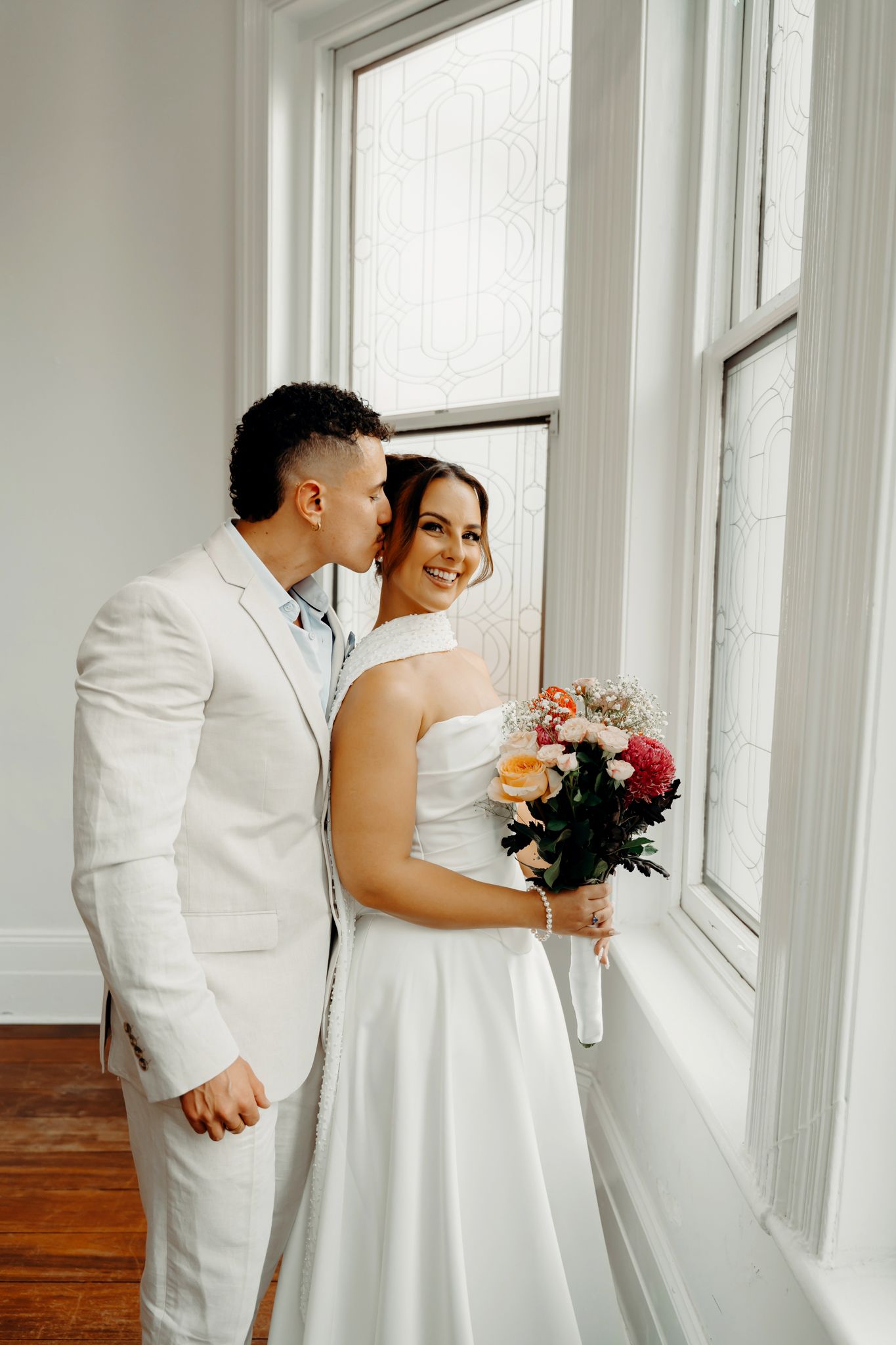 A smiling bride holding a colorful bouquet receives a kiss on the cheek from a groom in a white suit by a window.