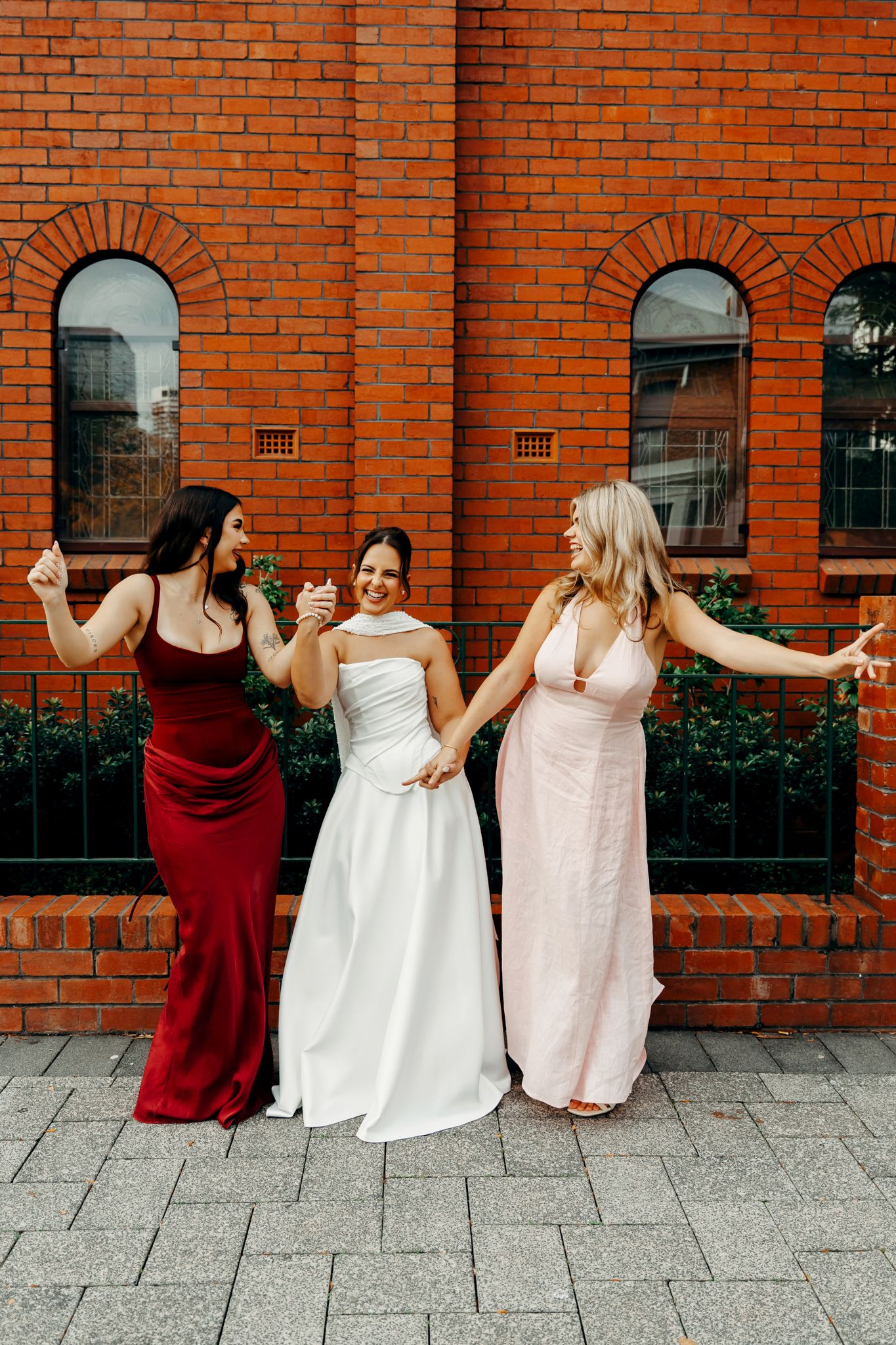 Bride in white gown celebrates with two bridesmaids in red and pink dresses outside a red brick building.