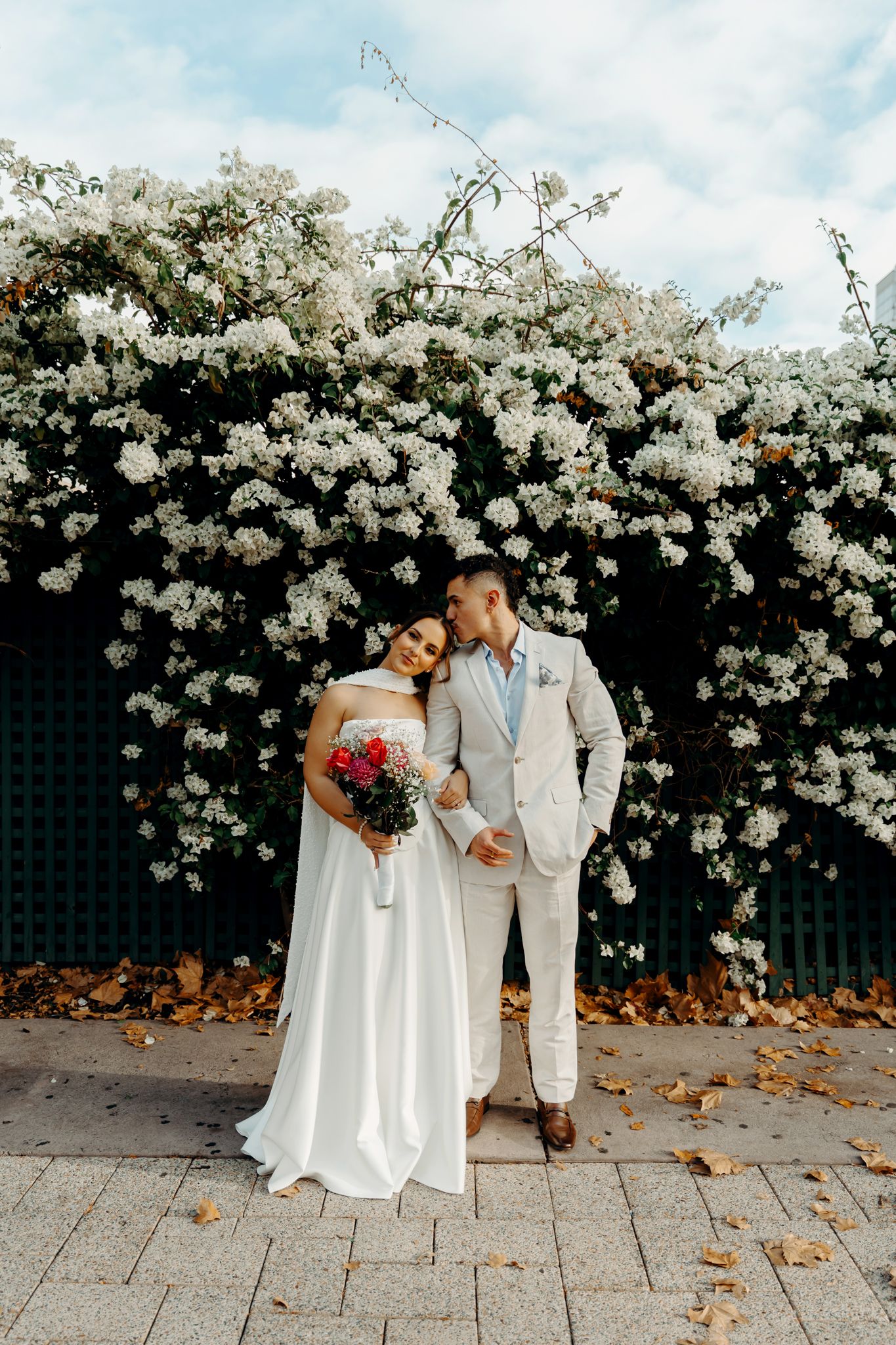 Bride and groom pose romantically before a lush white flowering hedge on their wedding day.
