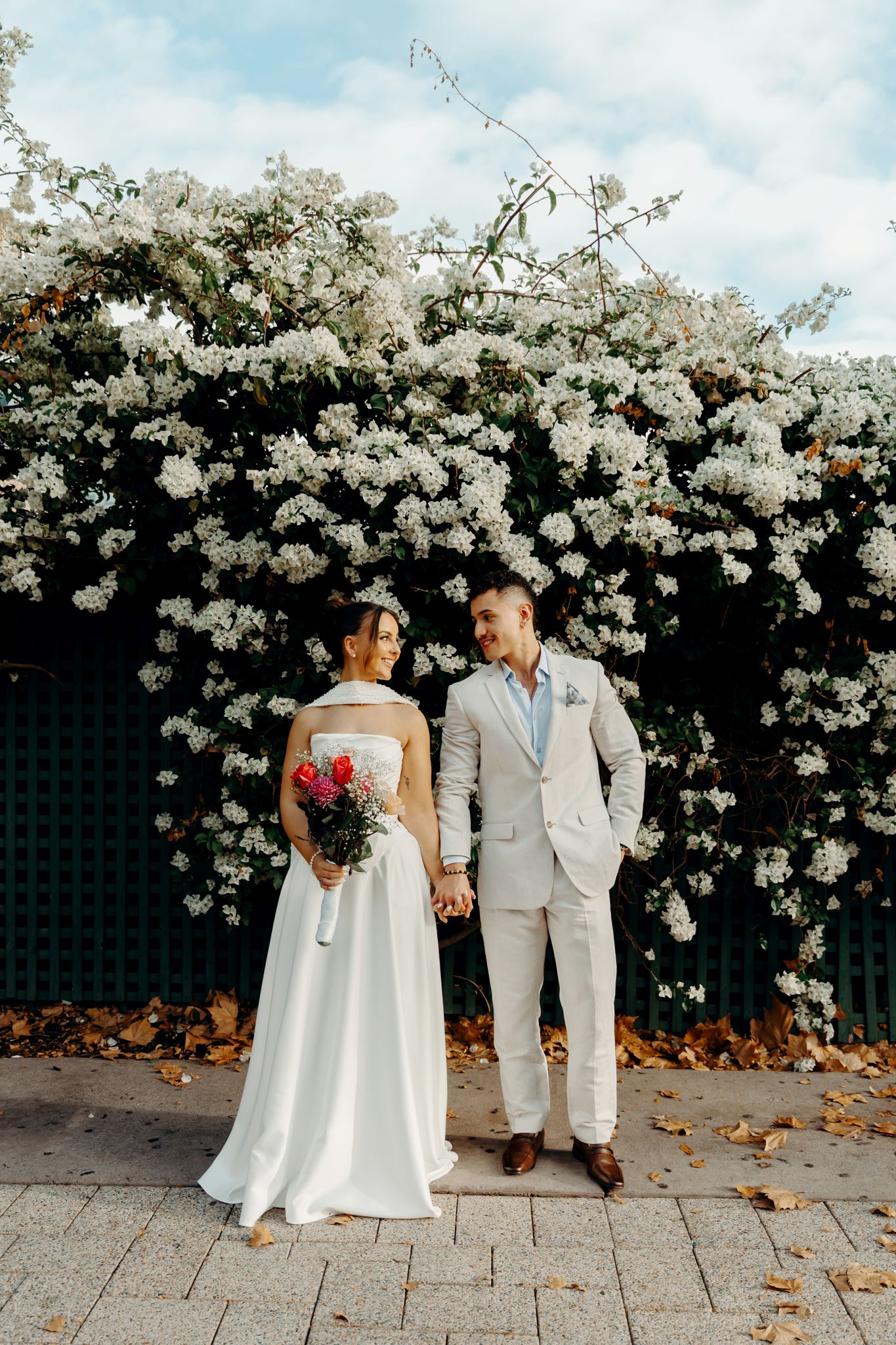 Bride and groom holding hands in front of blooming white flowers, bride holding red bouquet in white gown, groom in beige suit.