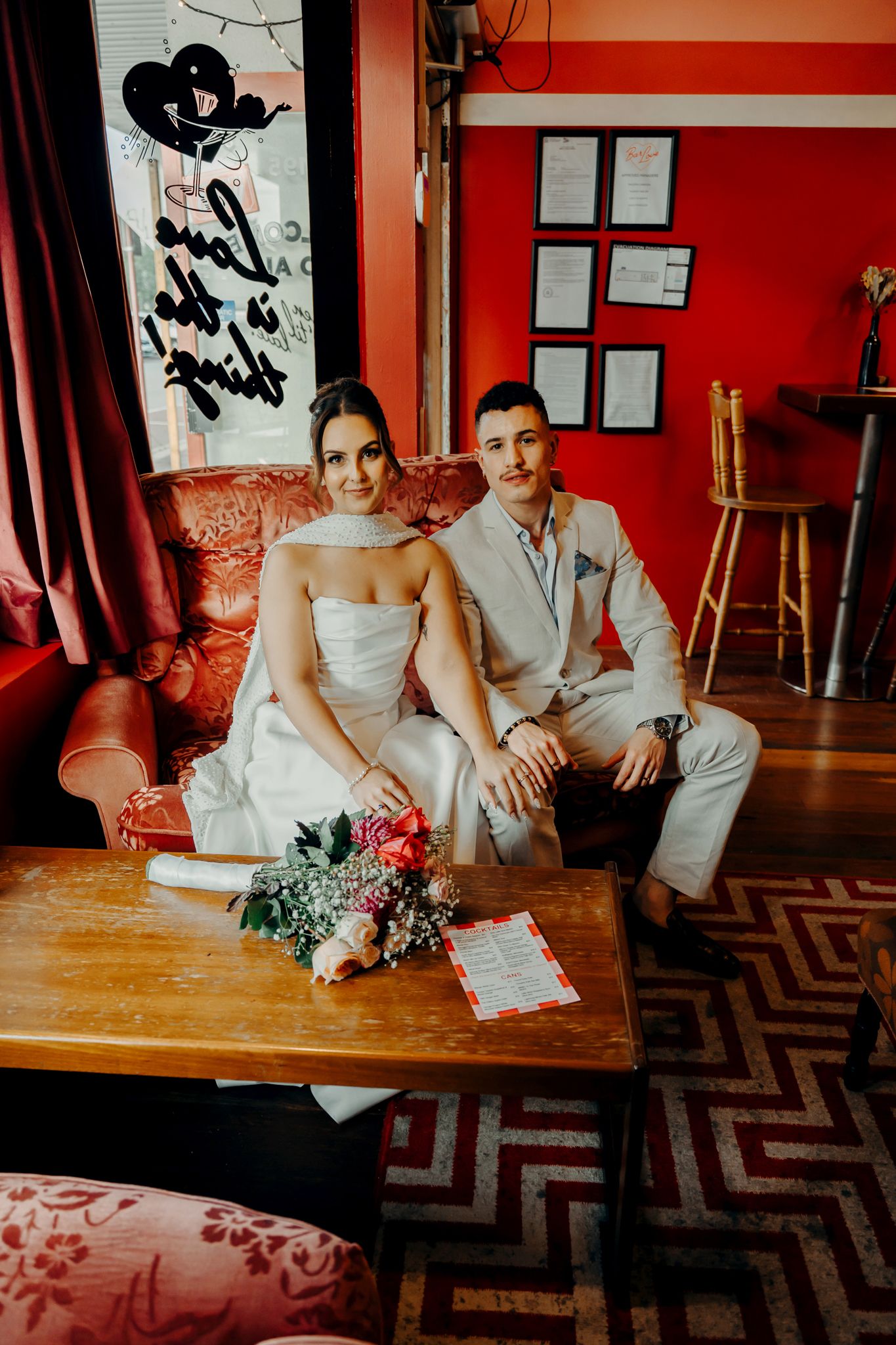 Bride and groom sitting together on a red sofa with a floral bouquet on the coffee table in a vibrant red interior.