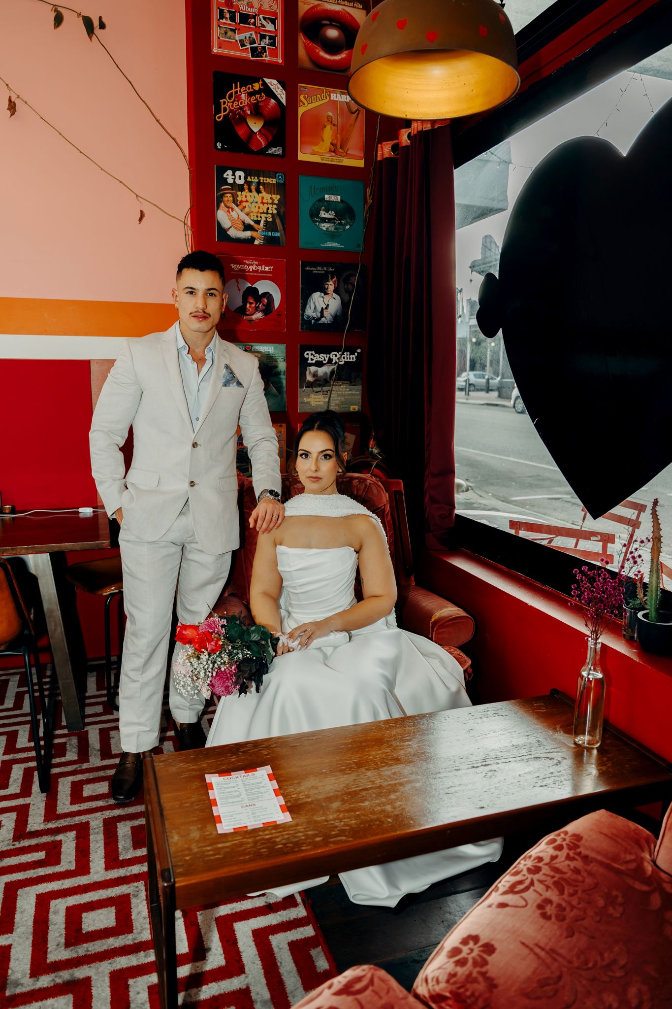 Bride and groom pose in a vibrant retro venue with vinyl records on red walls, colorful bouquet, and heart balloon.